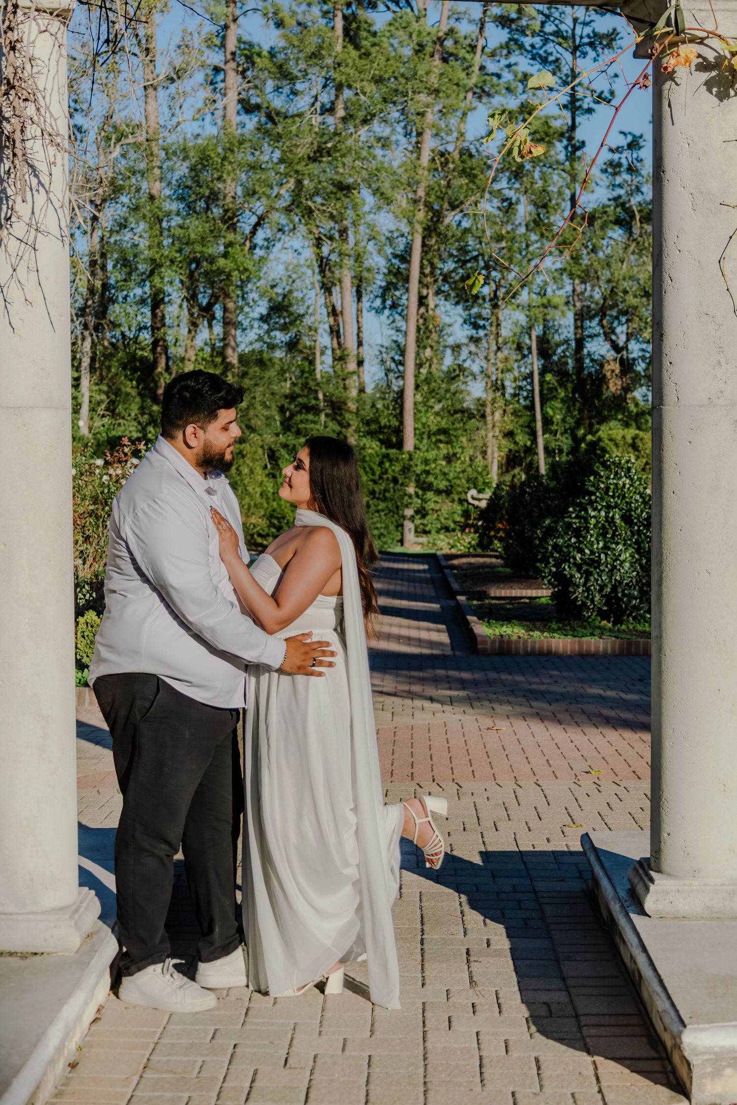 Engaged couple in white attire embracing under garden columns on brick pathway surrounded by lush trees.
