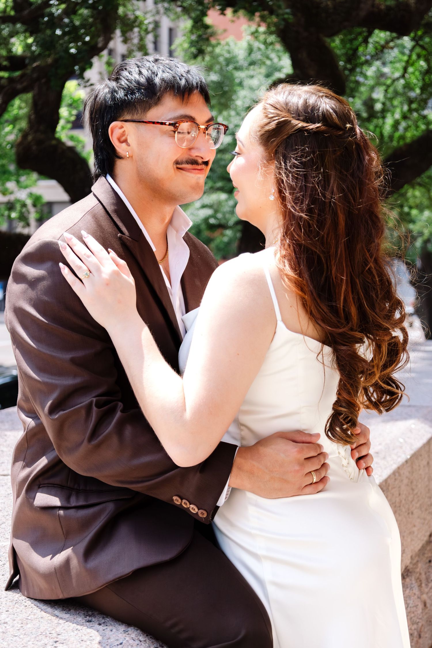 Bride and groom sitting on a stone ledge gazing at each other under lush green trees.