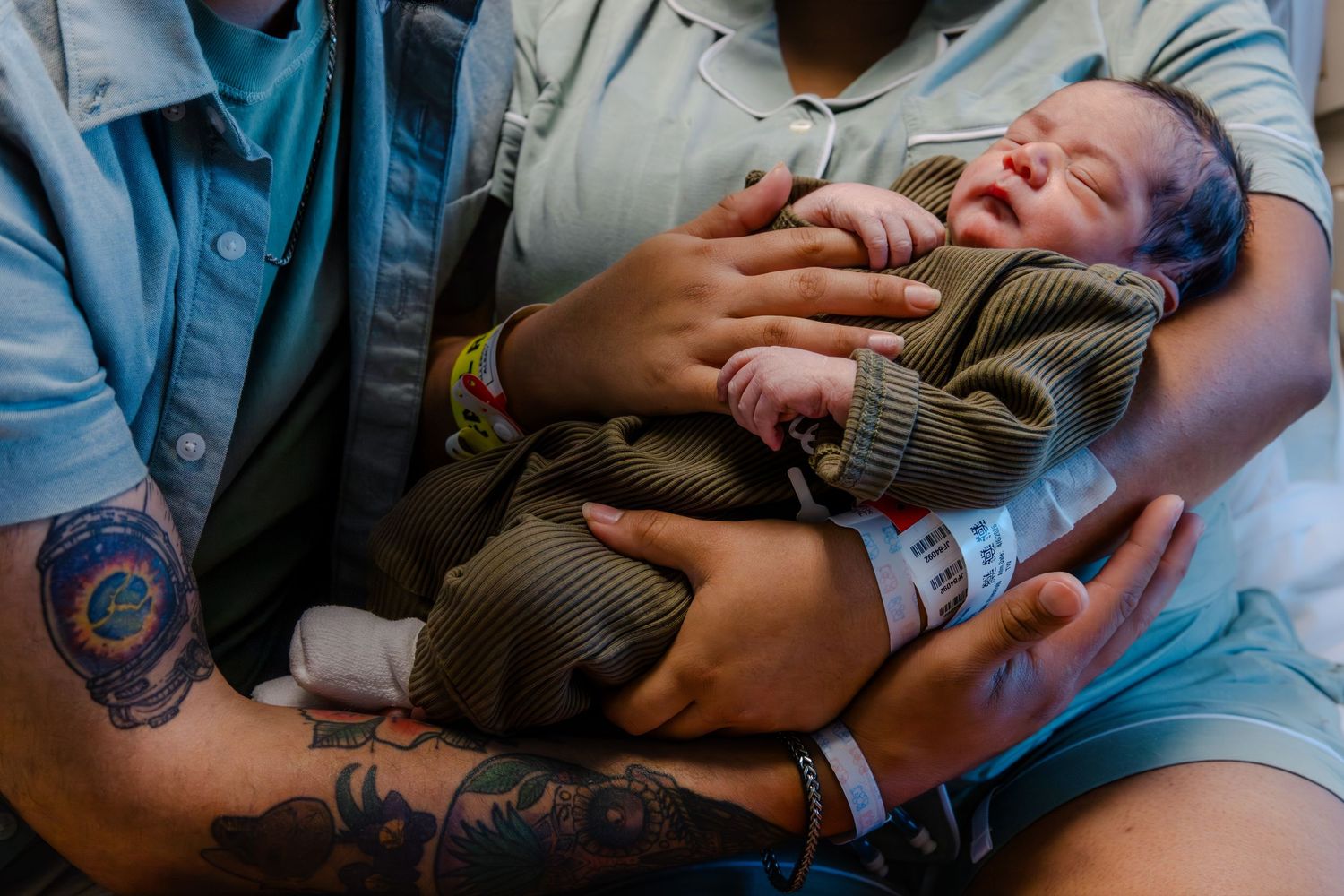 Parents lovingly hold their newborn baby wrapped in olive green, both wearing hospital ID bands.