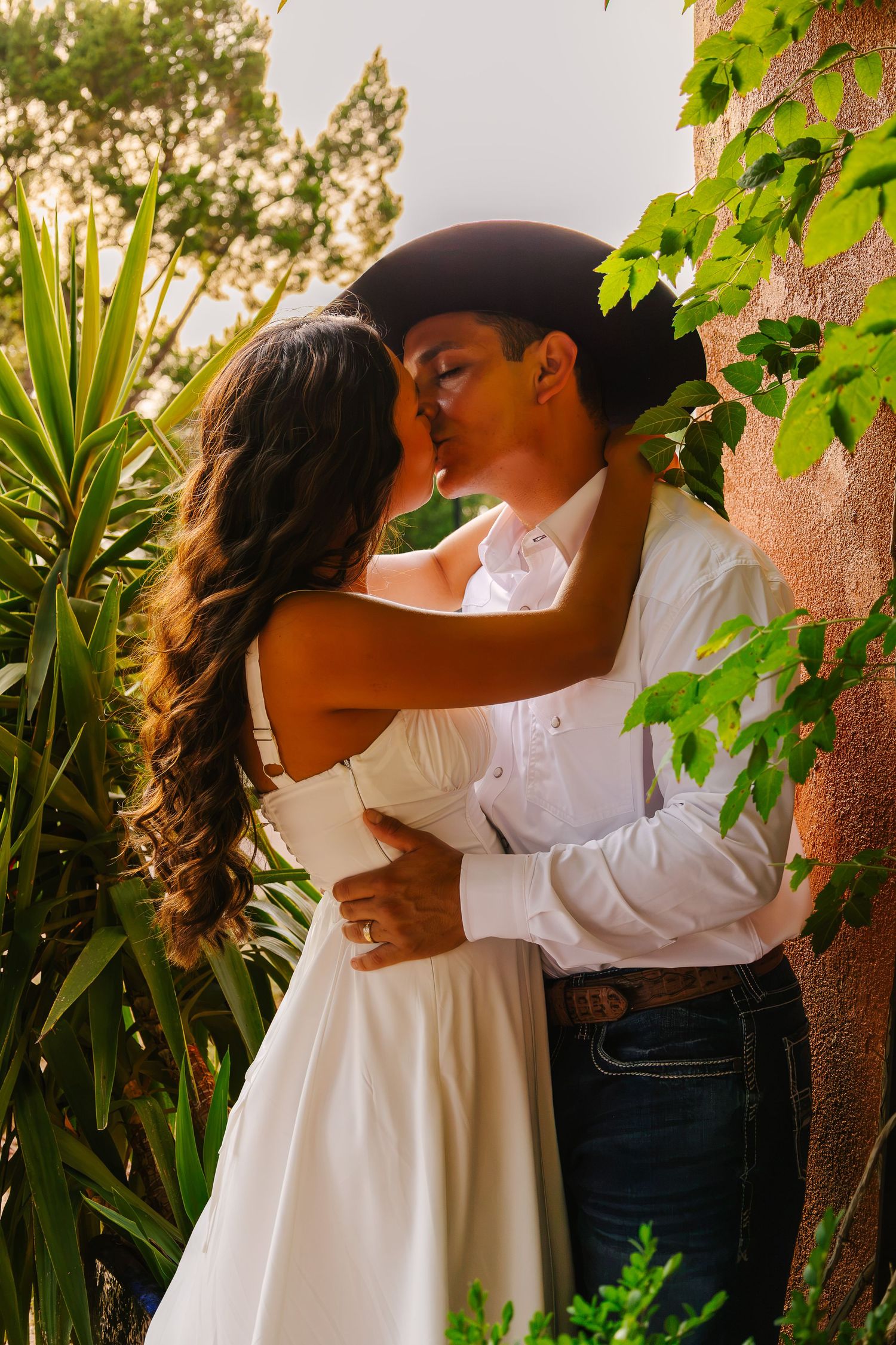 A couple shares a romantic kiss outdoors, man in cowboy hat and white shirt, woman in white dress, surrounded by lush greenery.