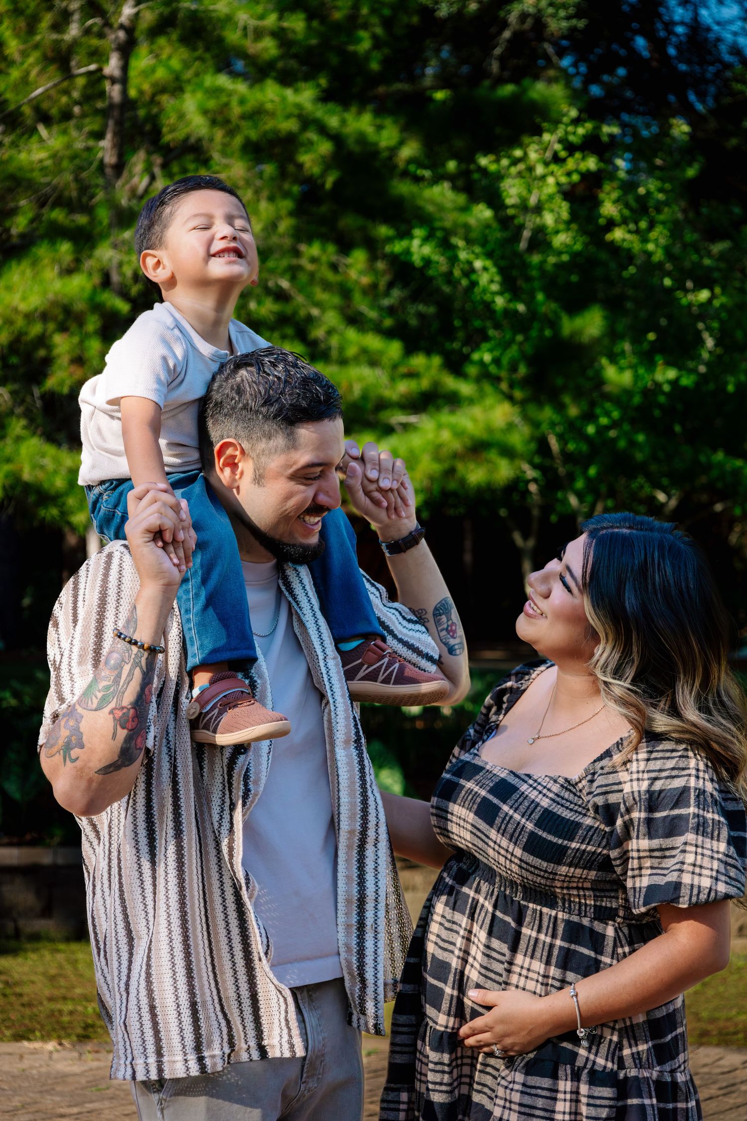 Joyful family outdoors with father carrying laughing toddler on shoulders while pregnant mother smiles lovingly.