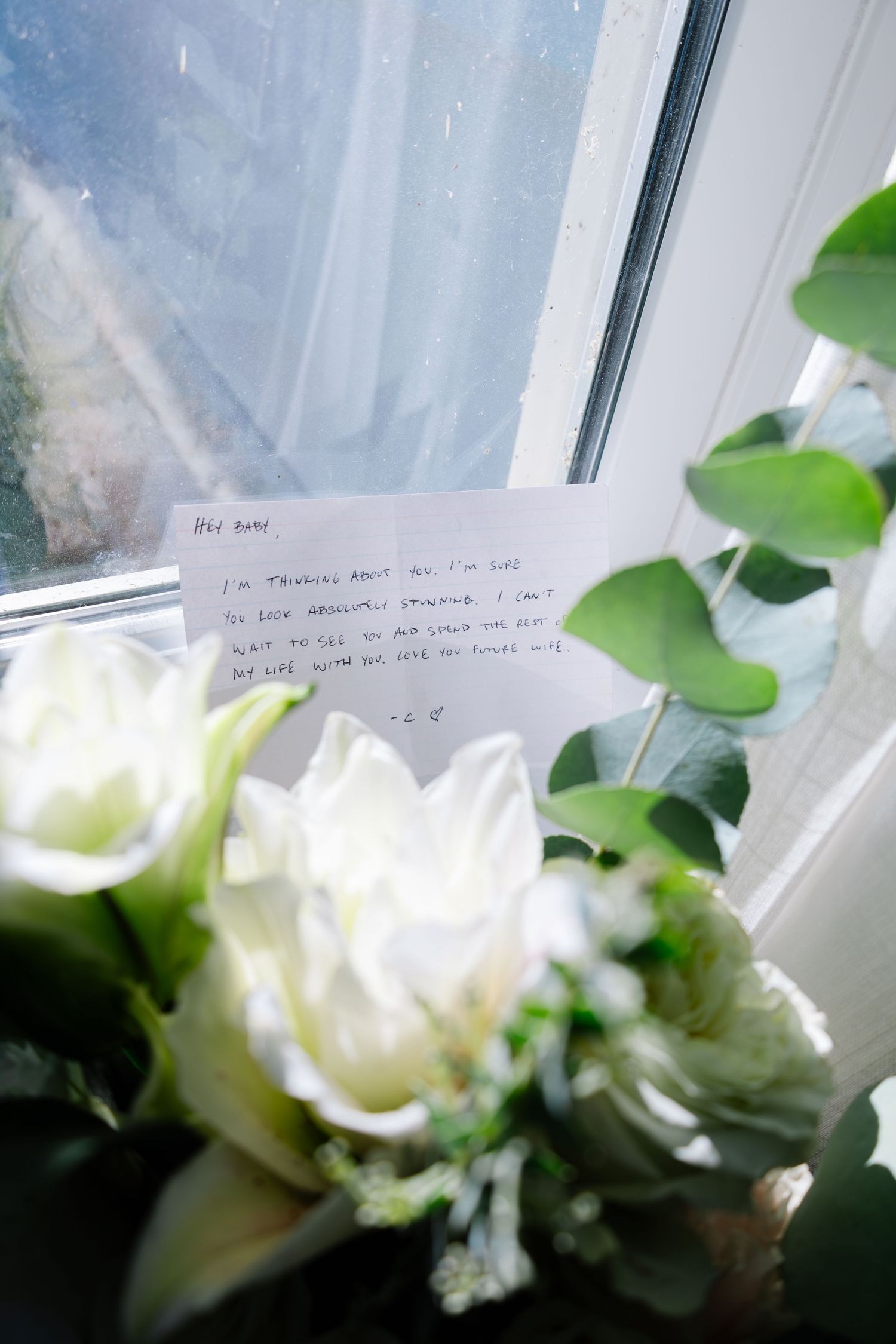 Handwritten wedding note placed beside white roses and eucalyptus bouquet on a windowsill.