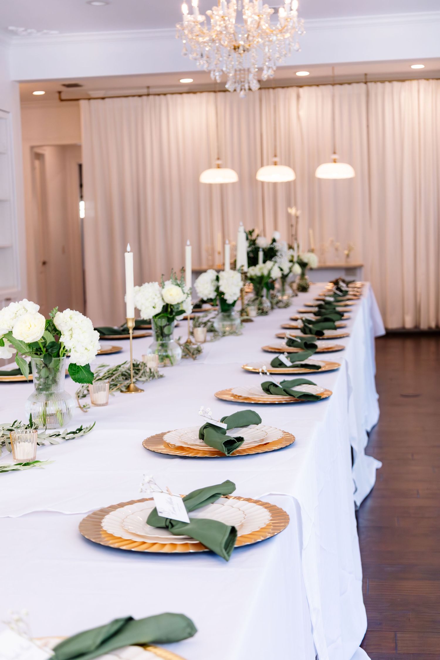 Elegant wedding reception table with white linens, gold chargers, green napkins, white floral centerpieces, and crystal chandelier.