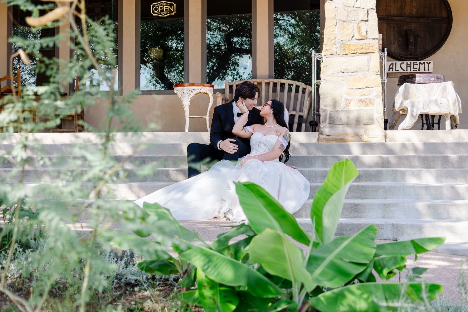 A bride and groom share a romantic moment seated on stone steps surrounded by lush greenery at an elegant venue.