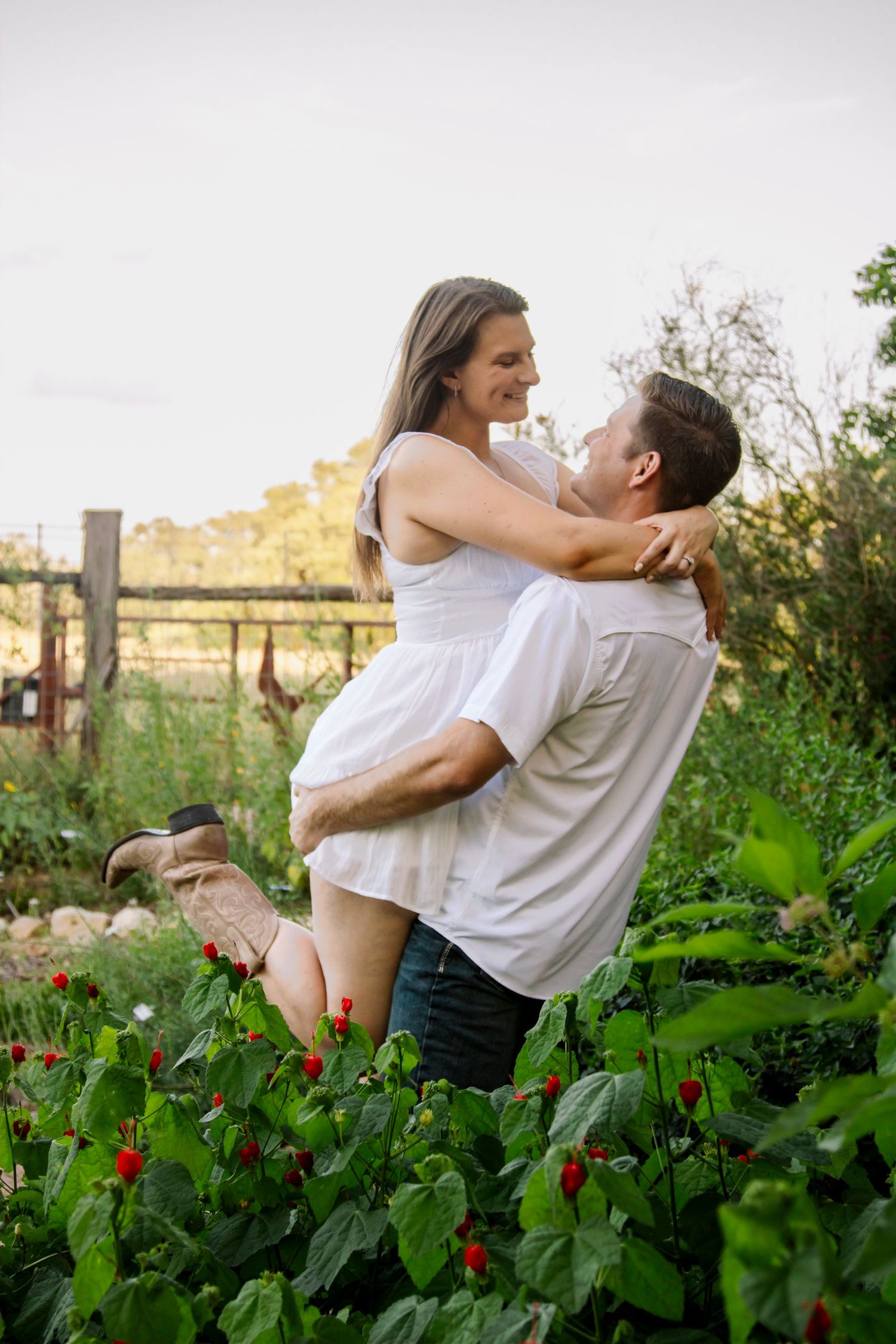 Man lifting woman in white dress and cowboy boots in a lush green garden during engagement photos.