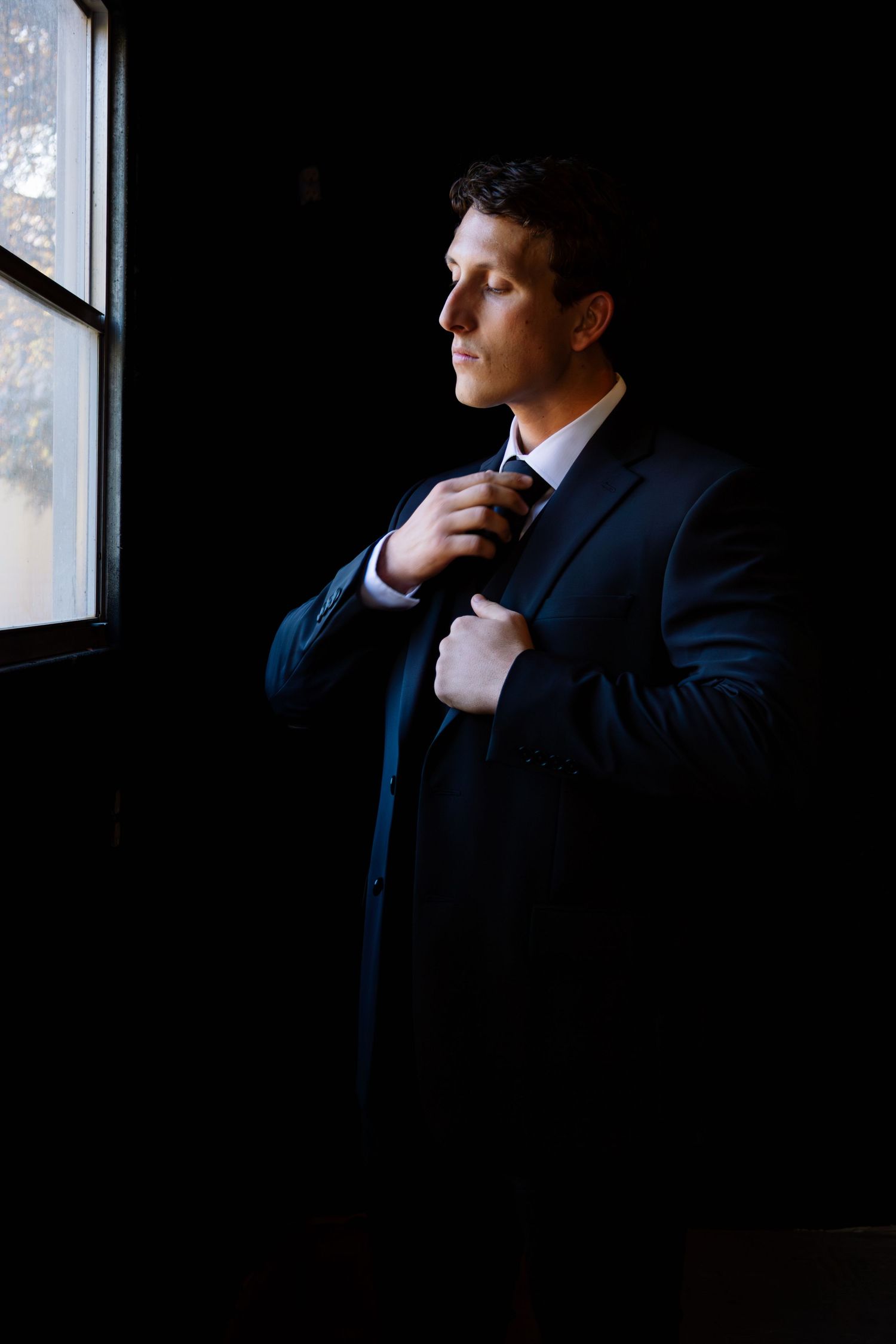 Groom in a dark suit adjusting his tie while standing by a window in dramatic lighting.