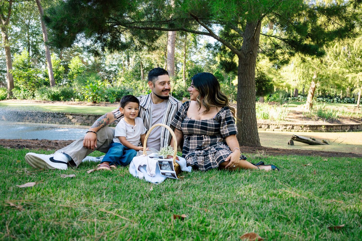 Happy family of three sits together on green grass in a sunny park, smiling warmly.