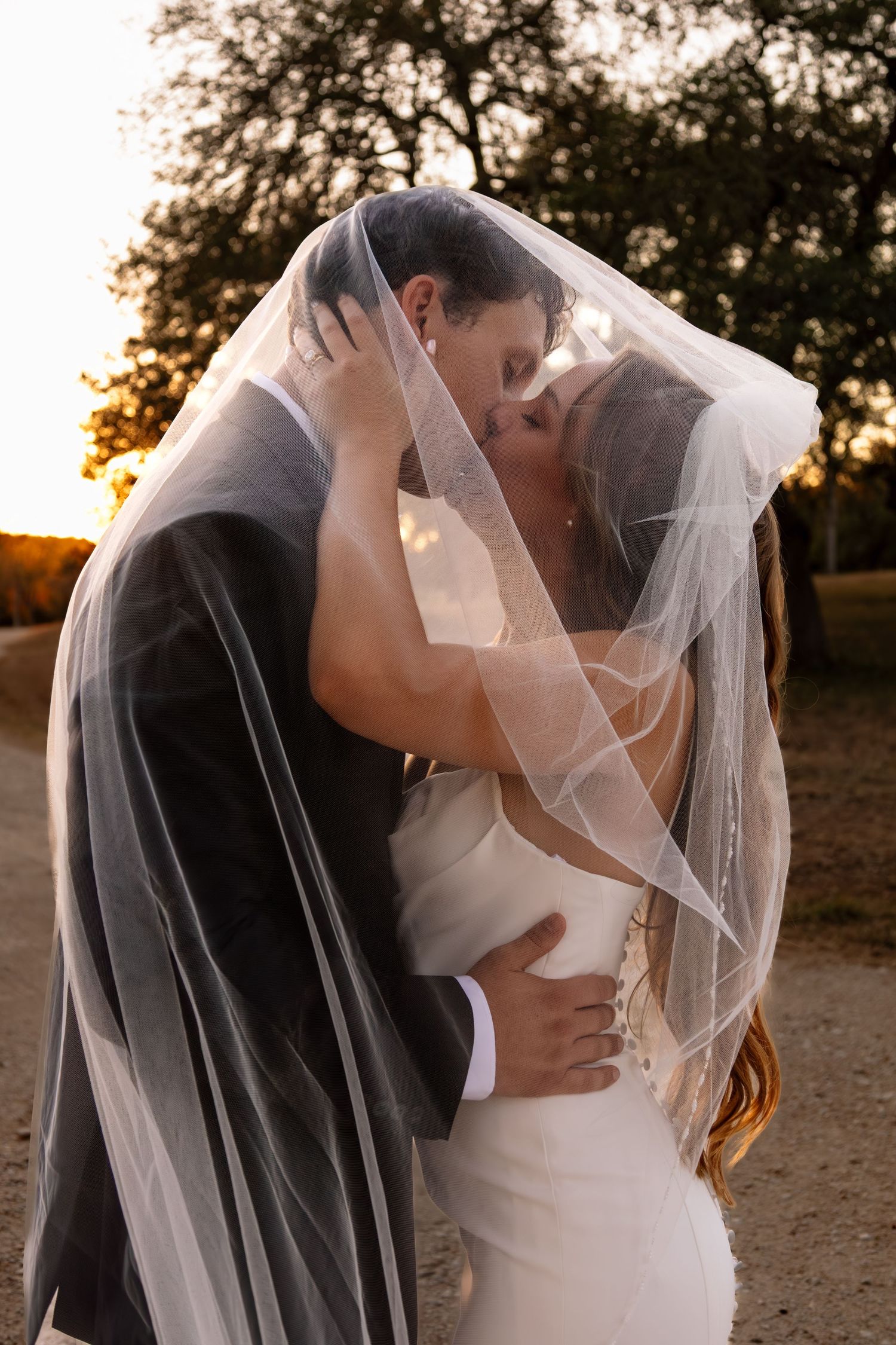 Bride and groom sharing a kiss under a wedding veil at golden hour with trees in the background.