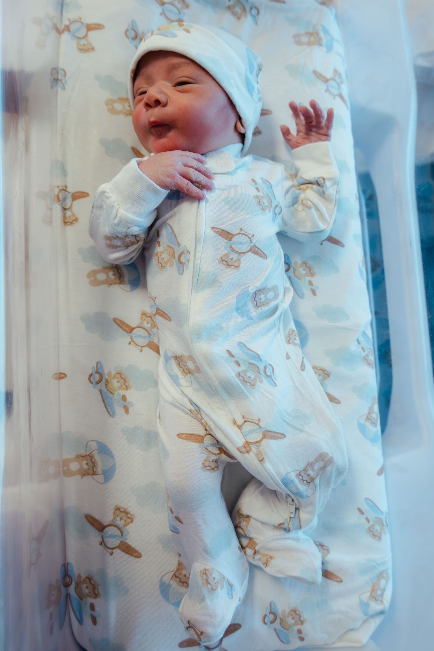 Newborn baby in airplane-print onesie and hat lying in a hospital bassinet with hand raised.