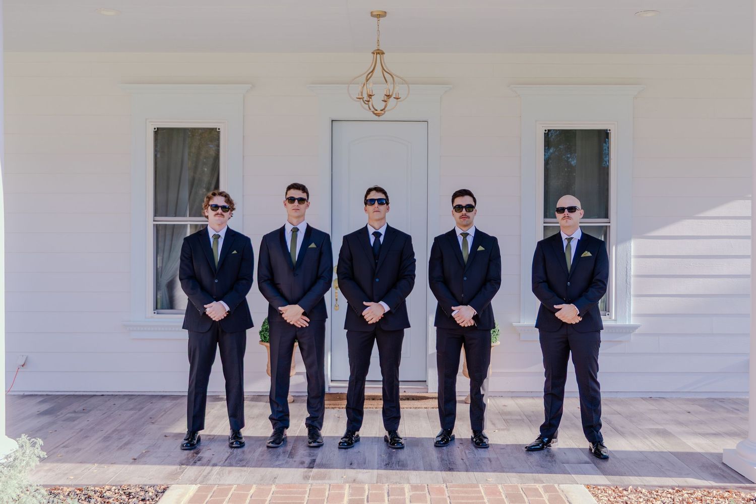 Five groomsmen in black suits and sunglasses posing in a row outside a white building.