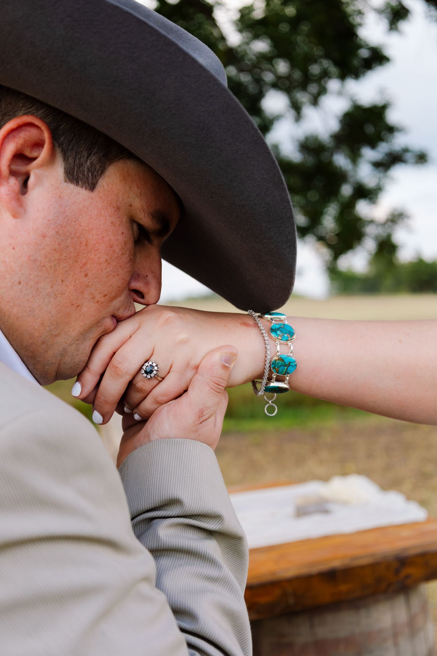 Groom in gray suit and cowboy hat kisses bride's hand adorned with turquoise bracelet and diamond ring.