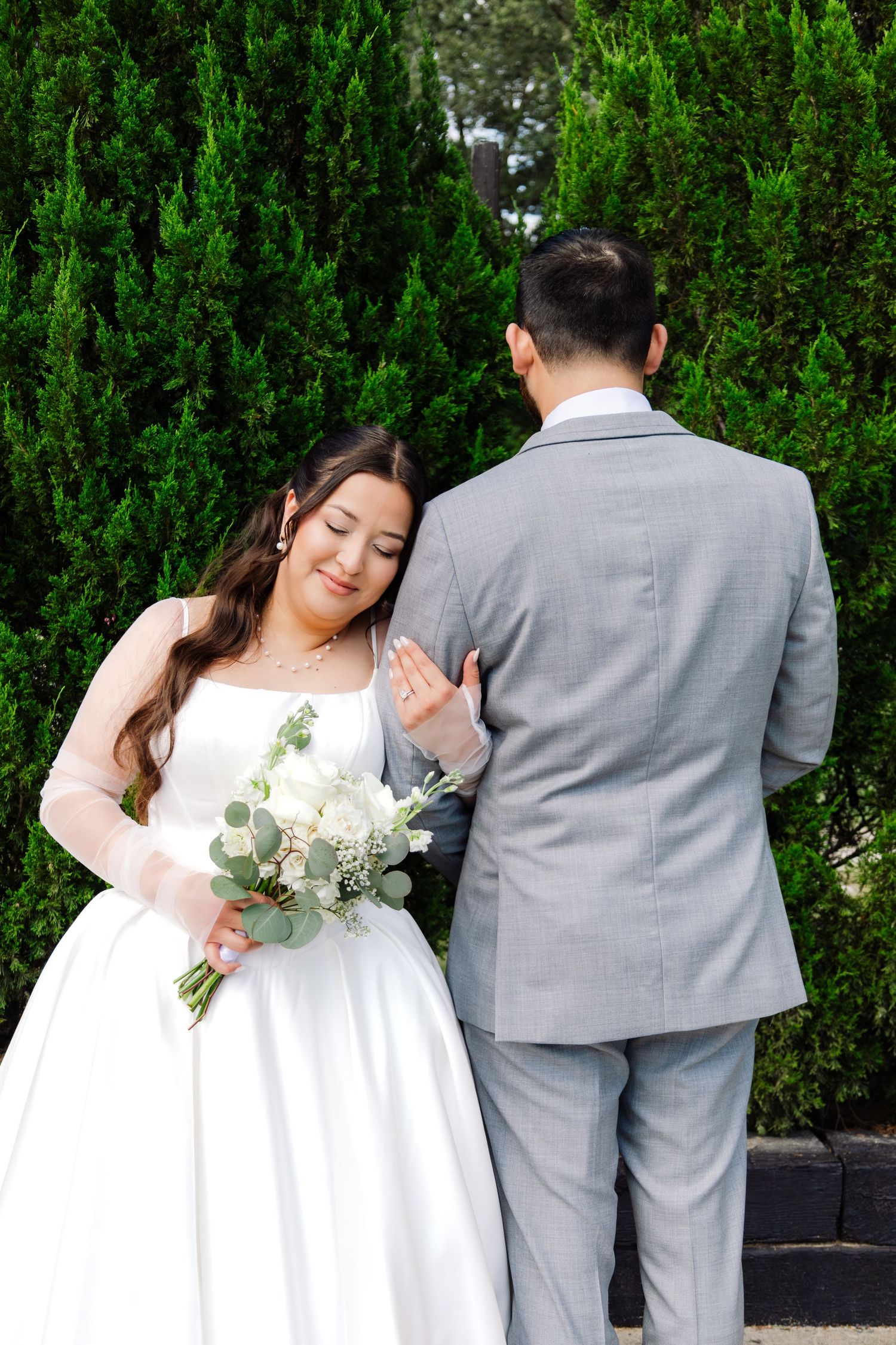 Bride in white gown holds bouquet of white roses and eucalyptus while groom in gray suit stands with his back turned.