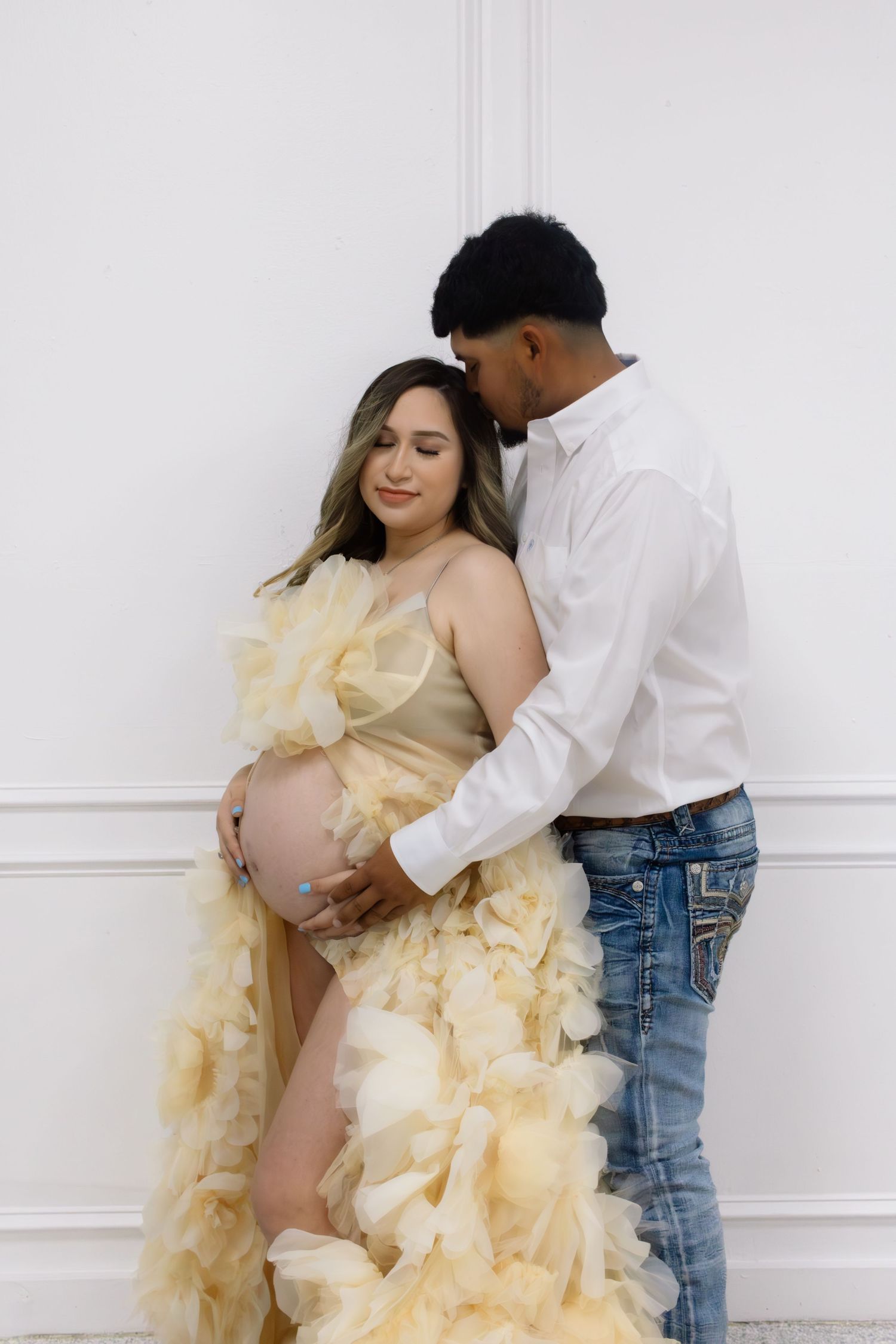 Pregnant woman in yellow ruffled gown holds her belly as her partner kisses her head by a white wall.
