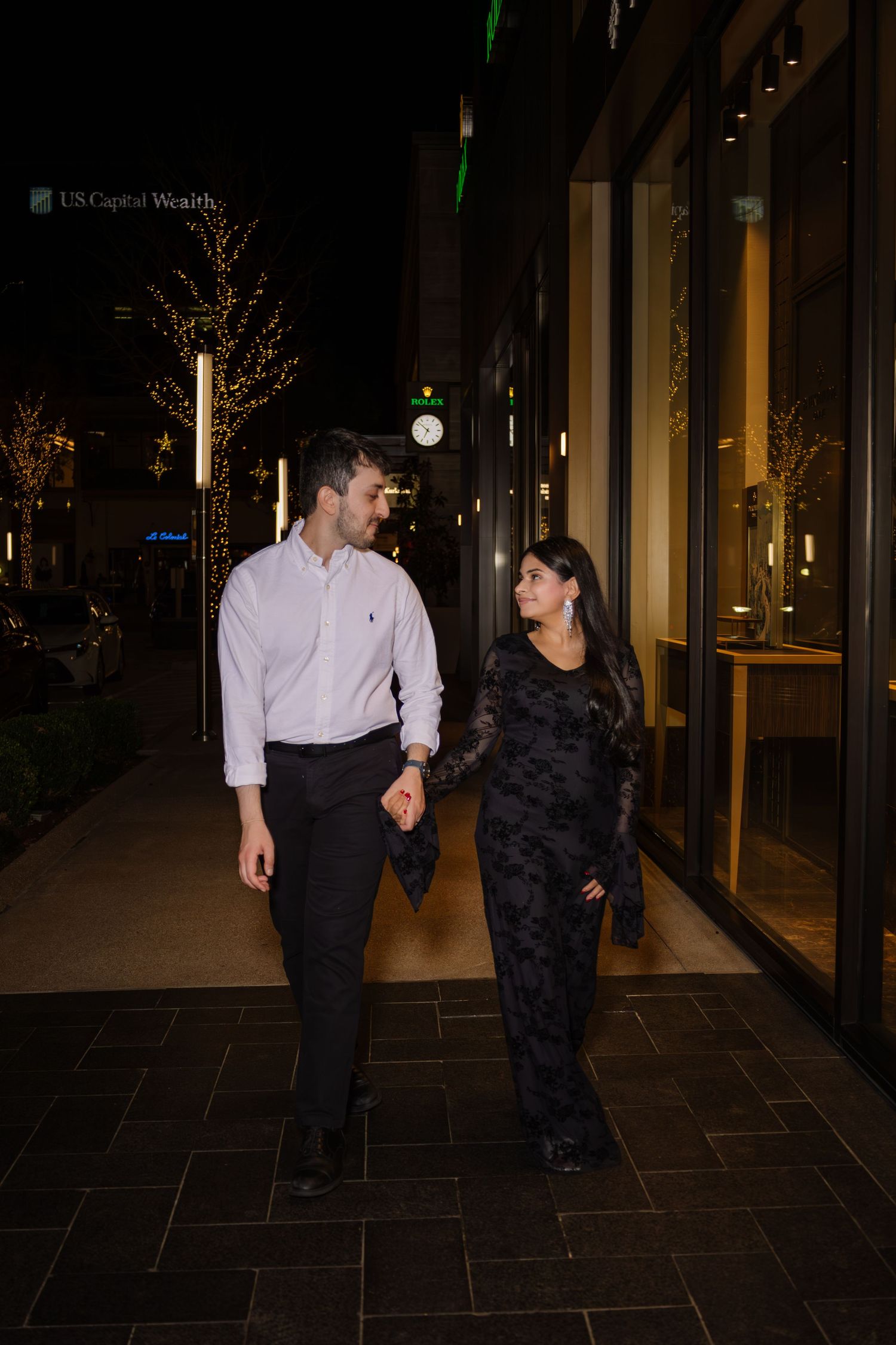 Engaged couple walking hand in hand on city sidewalk at night, woman in black dress and man in pink shirt.