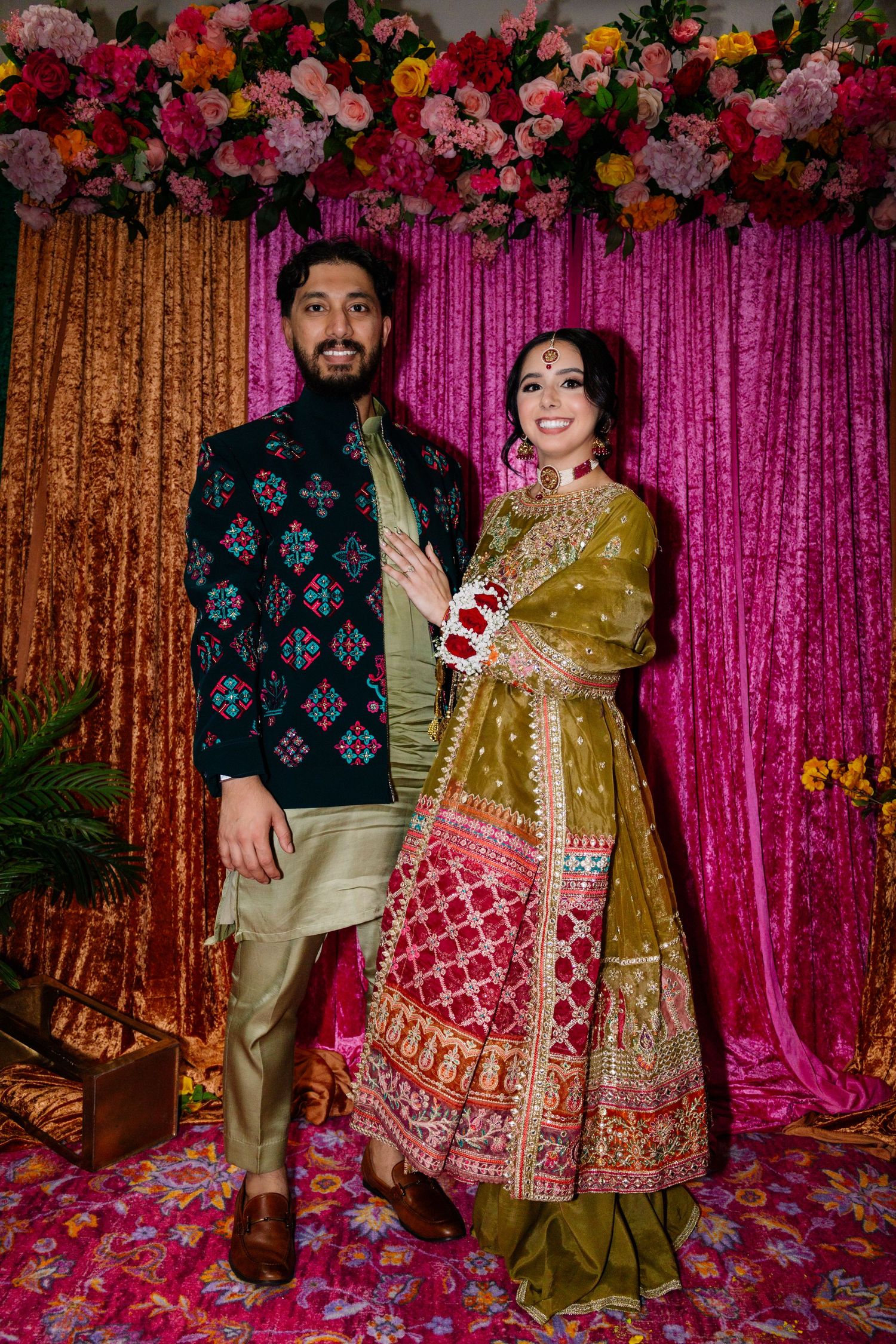 Indian bride and groom posing together in vibrant traditional wedding attire before a pink floral backdrop.