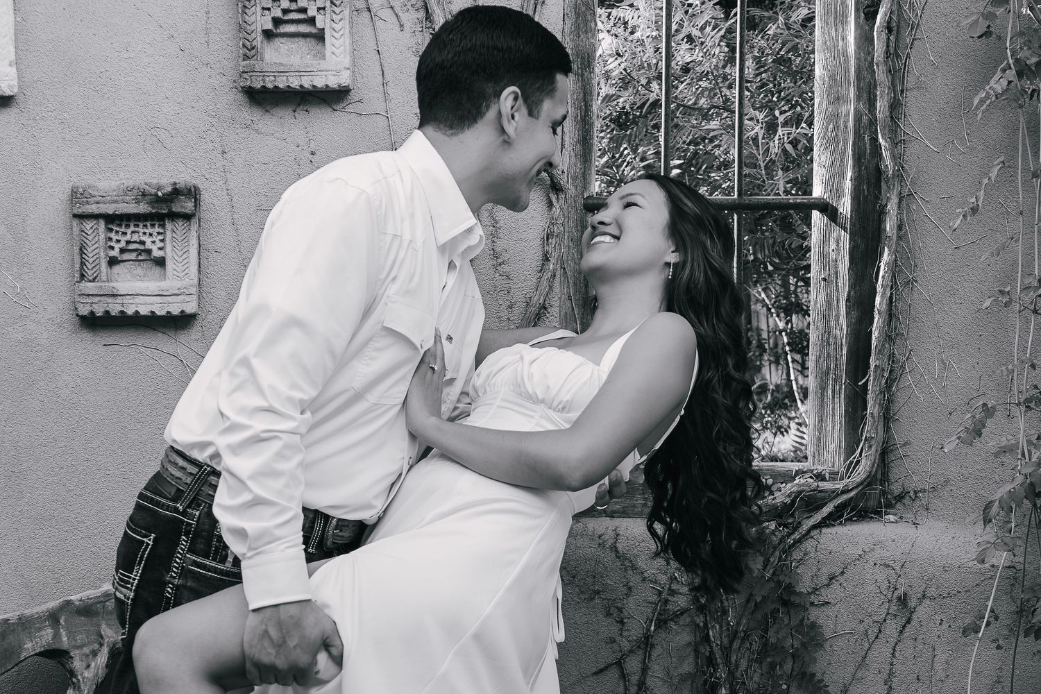 Black and white photo of a romantic couple sharing an intimate moment outdoors near a rustic wall and window.