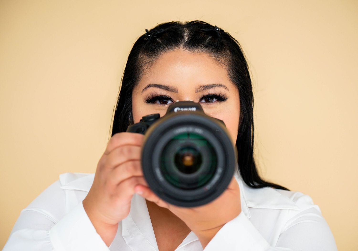 Woman photographer pointing a Canon DSLR camera directly at viewer against a beige background.