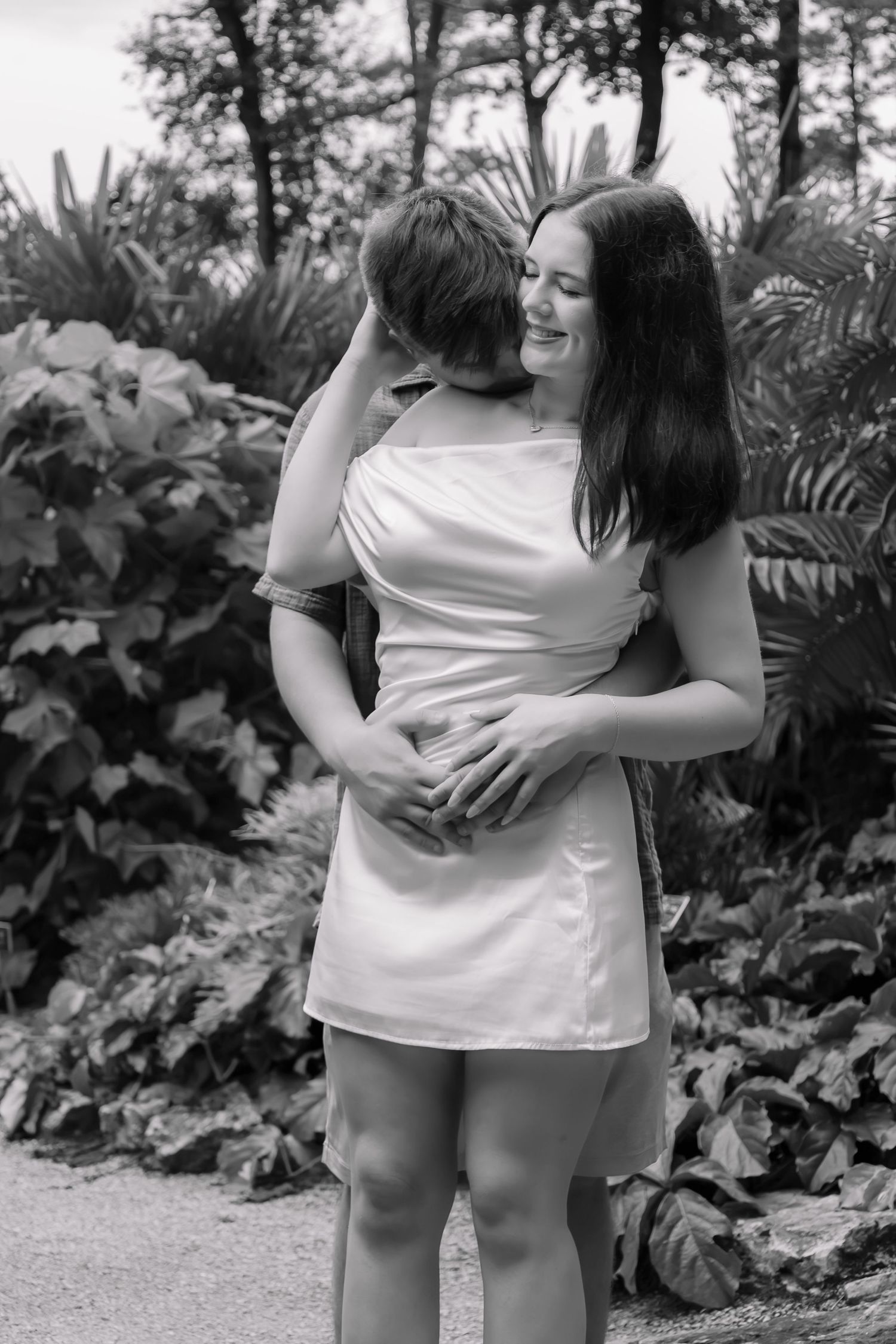 Black and white photo of a couple embracing in a lush garden, woman in white dress smiling joyfully.