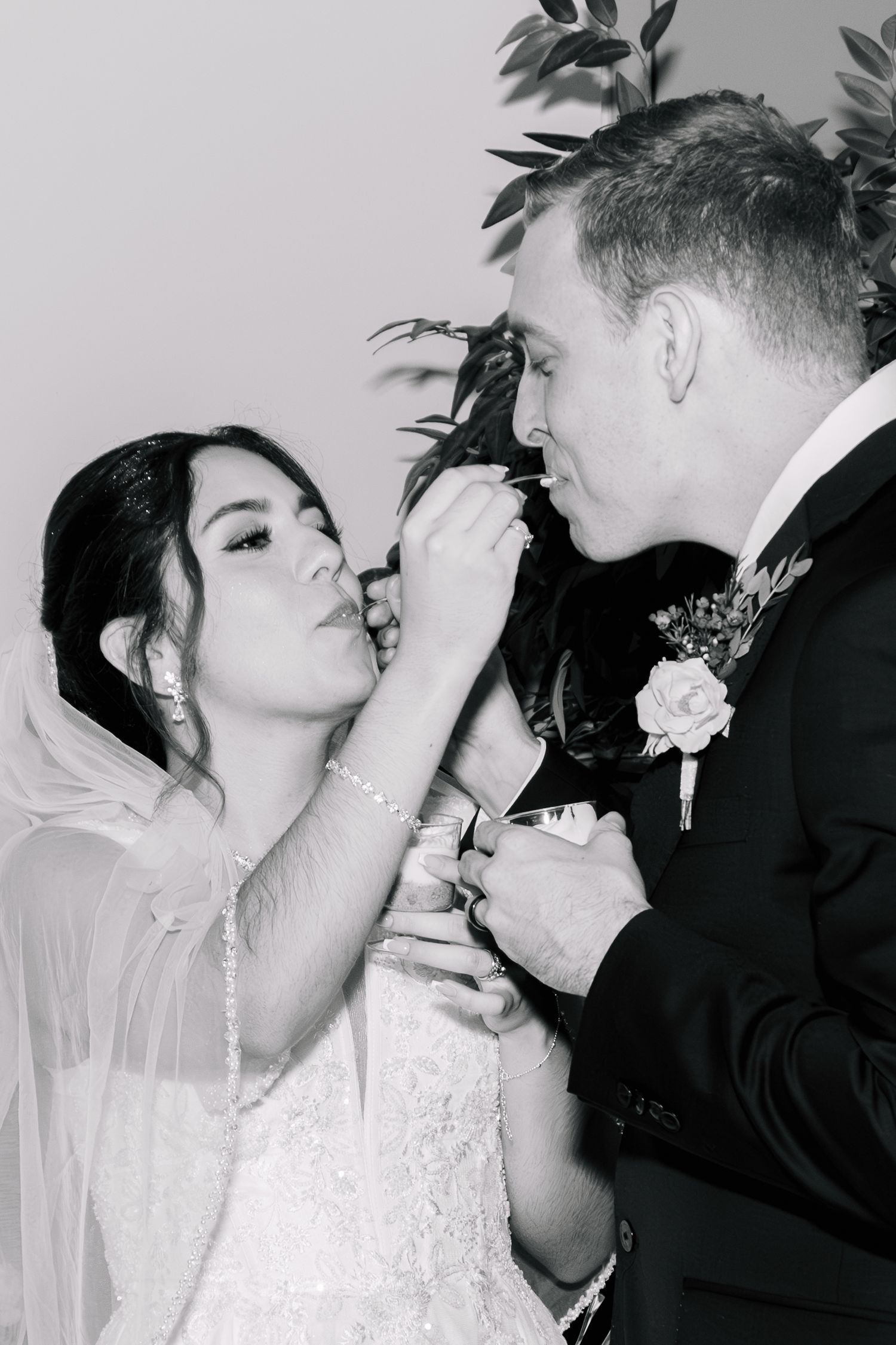 Black and white photo of a bride kissing the groom's hand near a floral boutonniere indoors.