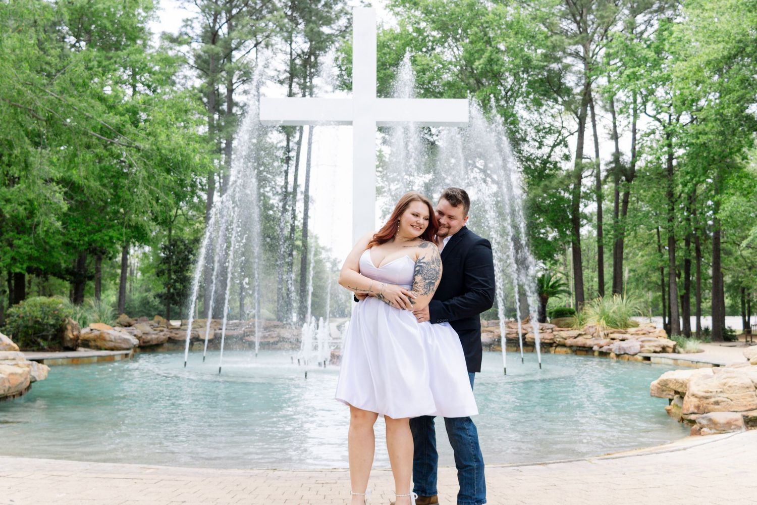 Couple embracing and smiling in front of a cross fountain surrounded by lush green trees outdoors.