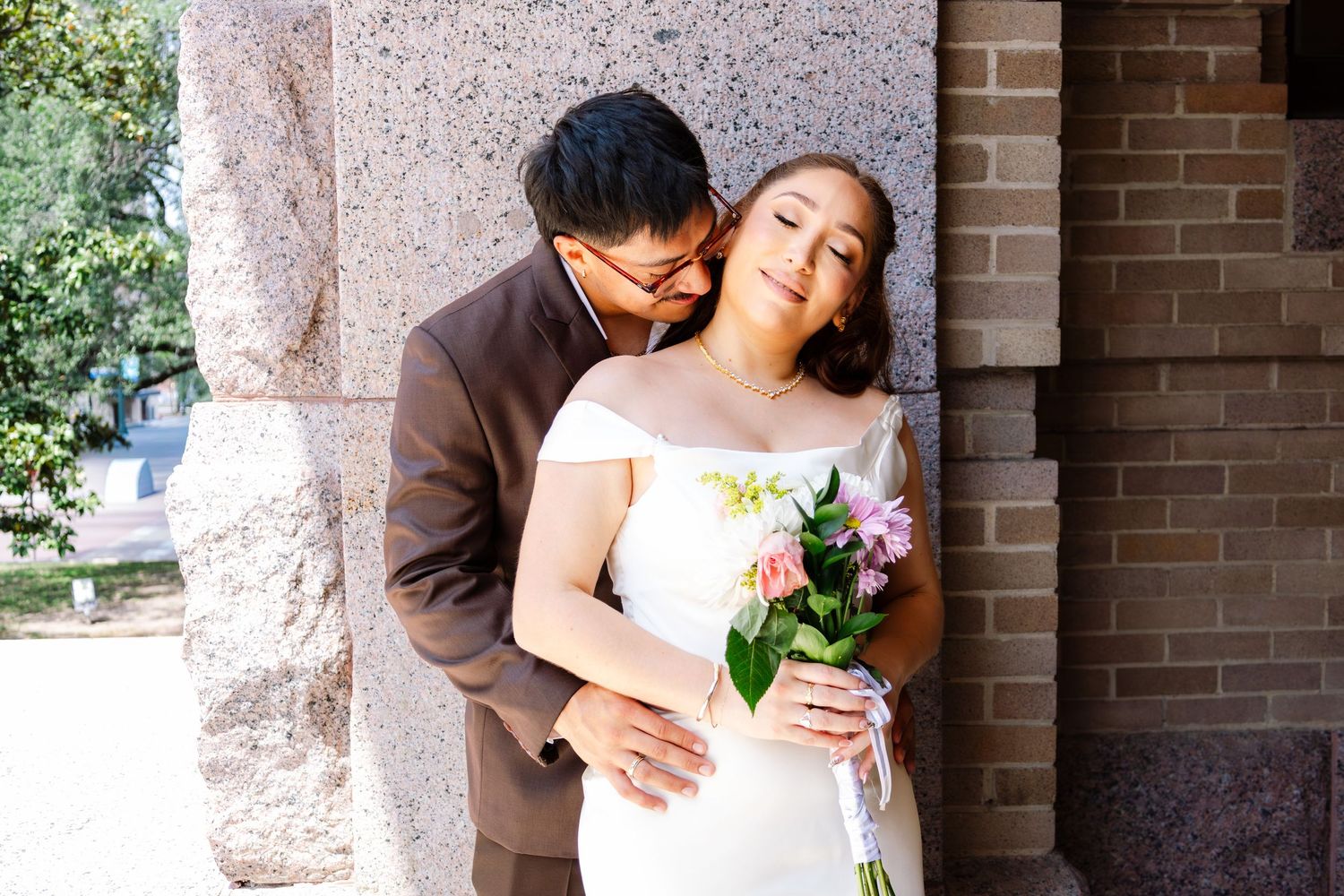 Groom tenderly kisses bride on cheek as she smiles holding a pink floral bouquet.