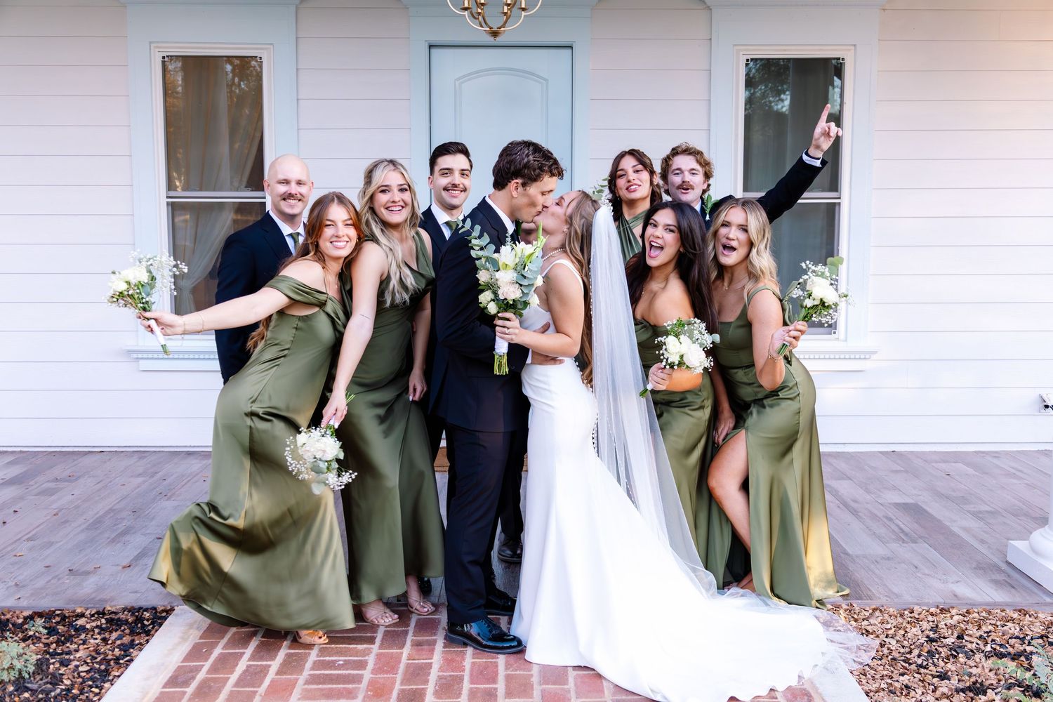 Wedding party celebrating outside white house with bride in veil, groom, and bridesmaids in olive green dresses.