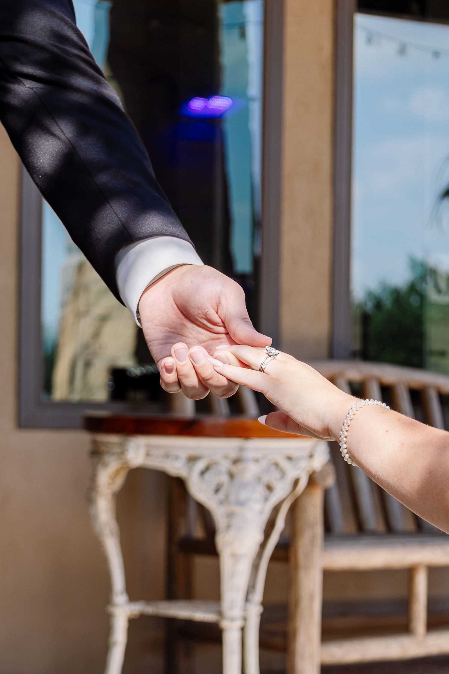 Bride and groom holding hands, showcasing wedding ring and pearl bracelet, with elegant white furniture in background.