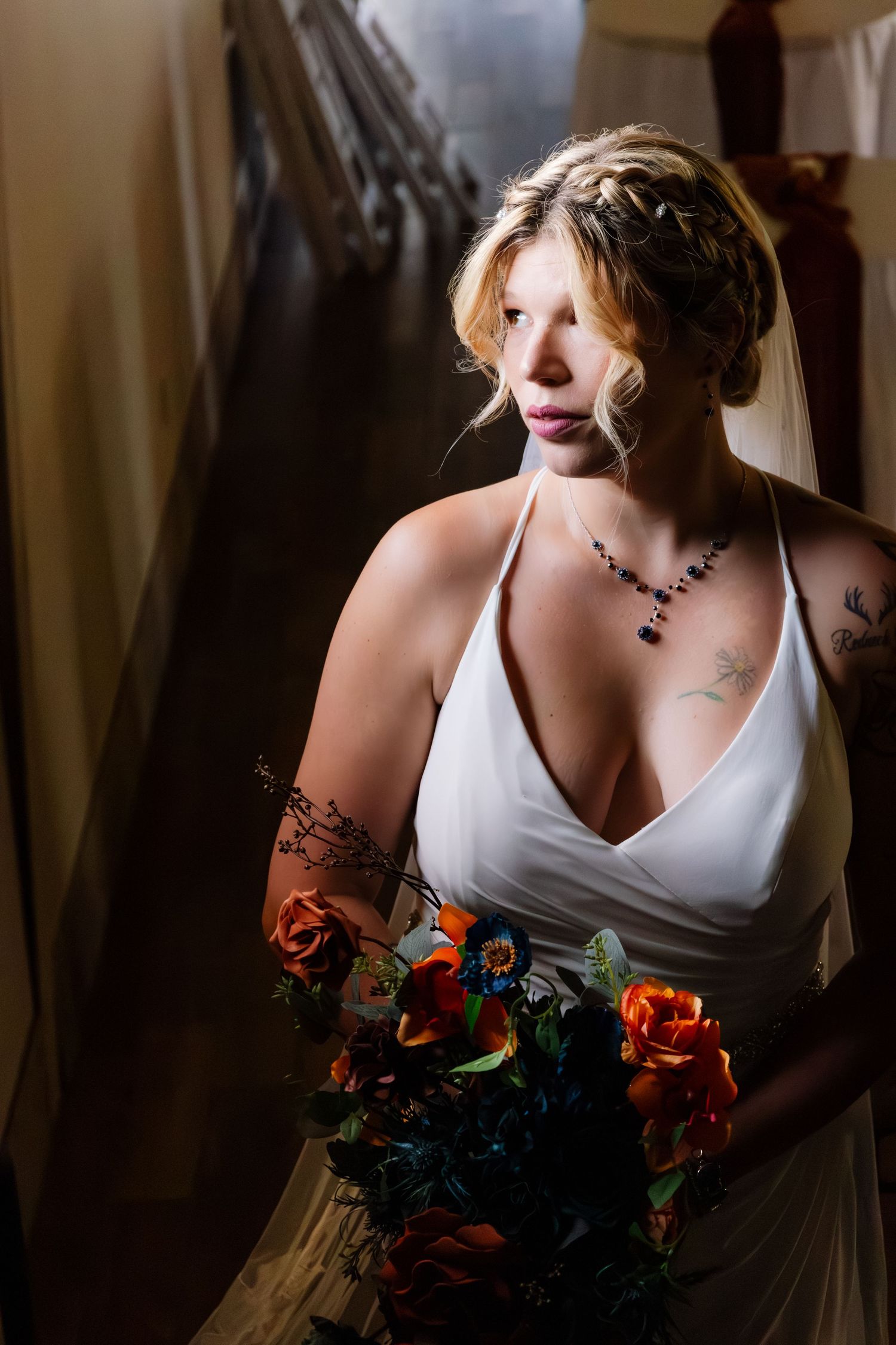 Bride in a white halter gown holds a vibrant orange and blue bouquet while standing in dramatic lighting.