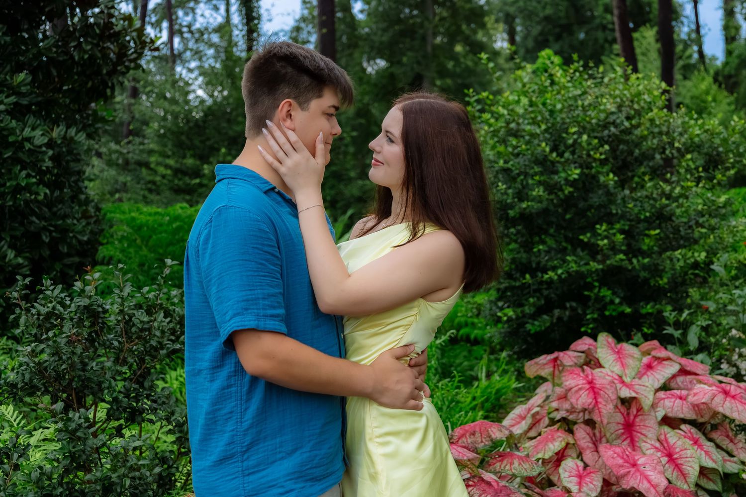 Young couple embracing romantically in a lush green garden with pink caladium plants, woman in yellow dress touching mans face.