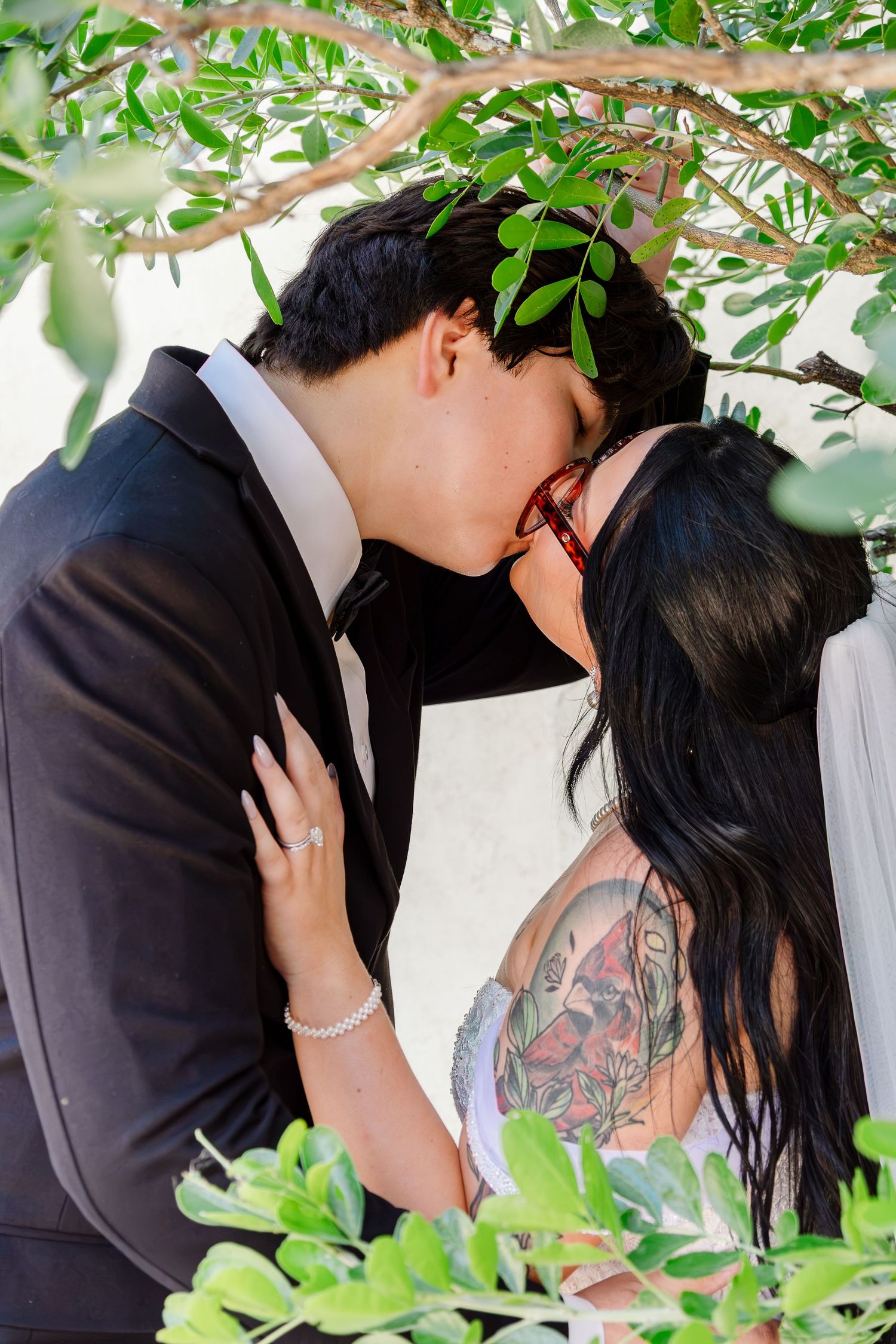Bride and groom sharing a first kiss under green tree branches, bride wearing white dress with floral tattoo sleeve.