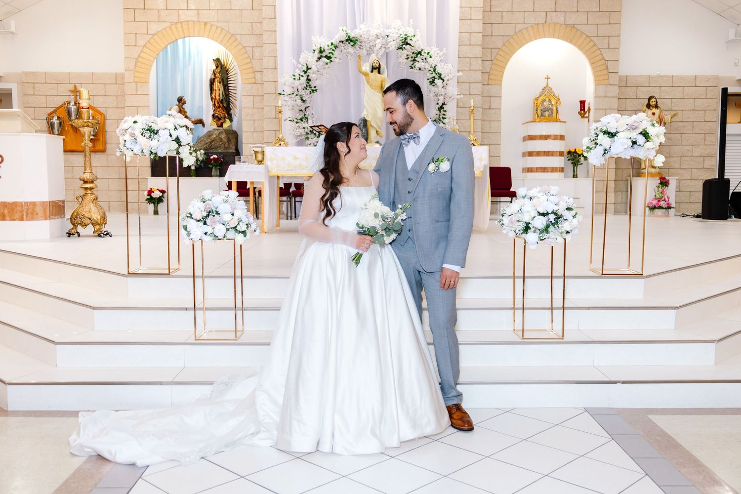 Bride and groom pose together at elegant white church altar adorned with gold stands and white floral arrangements.