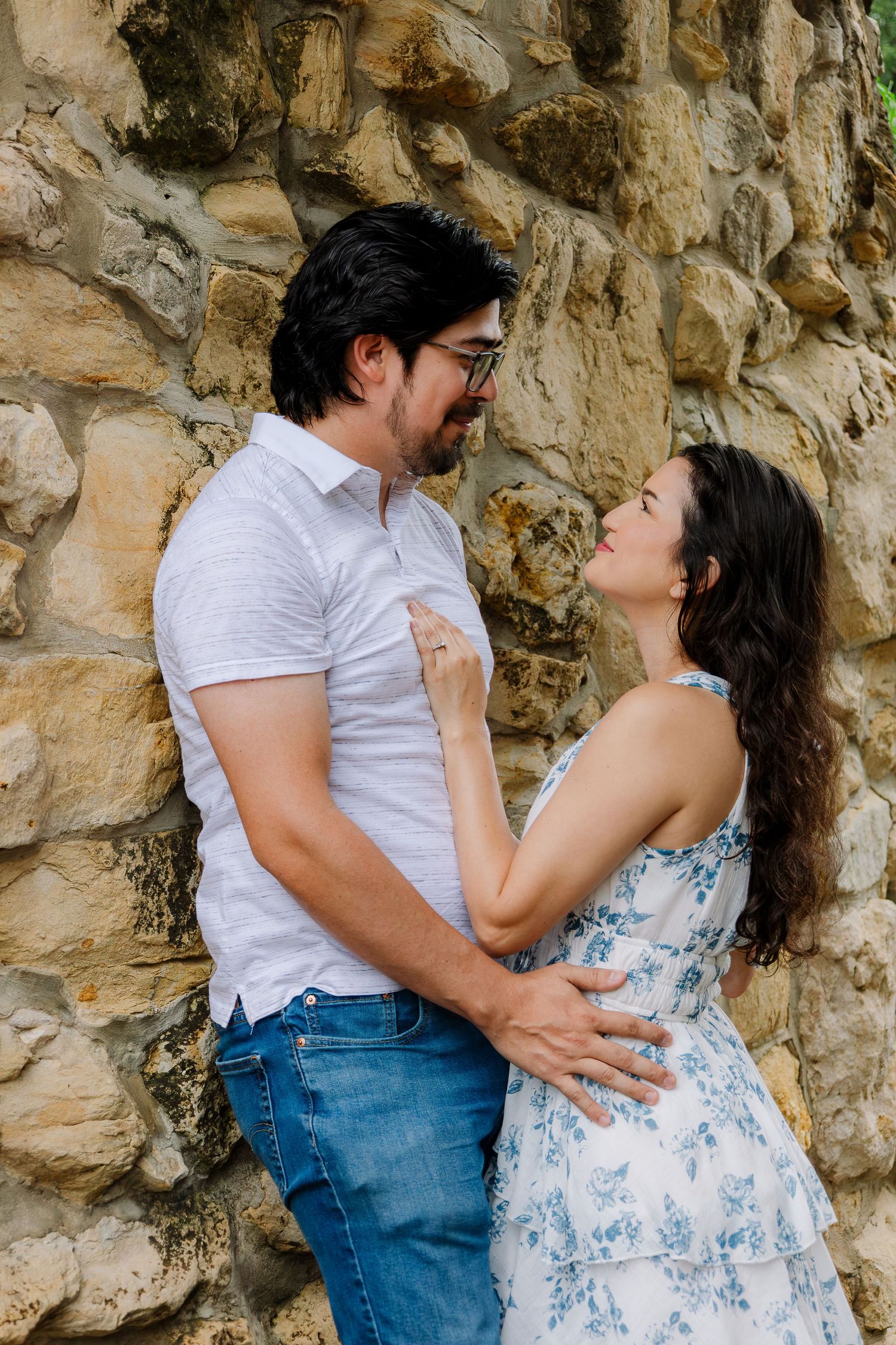 Couple embracing and gazing at each other against a rustic stone wall, man in white shirt and woman in floral dress.