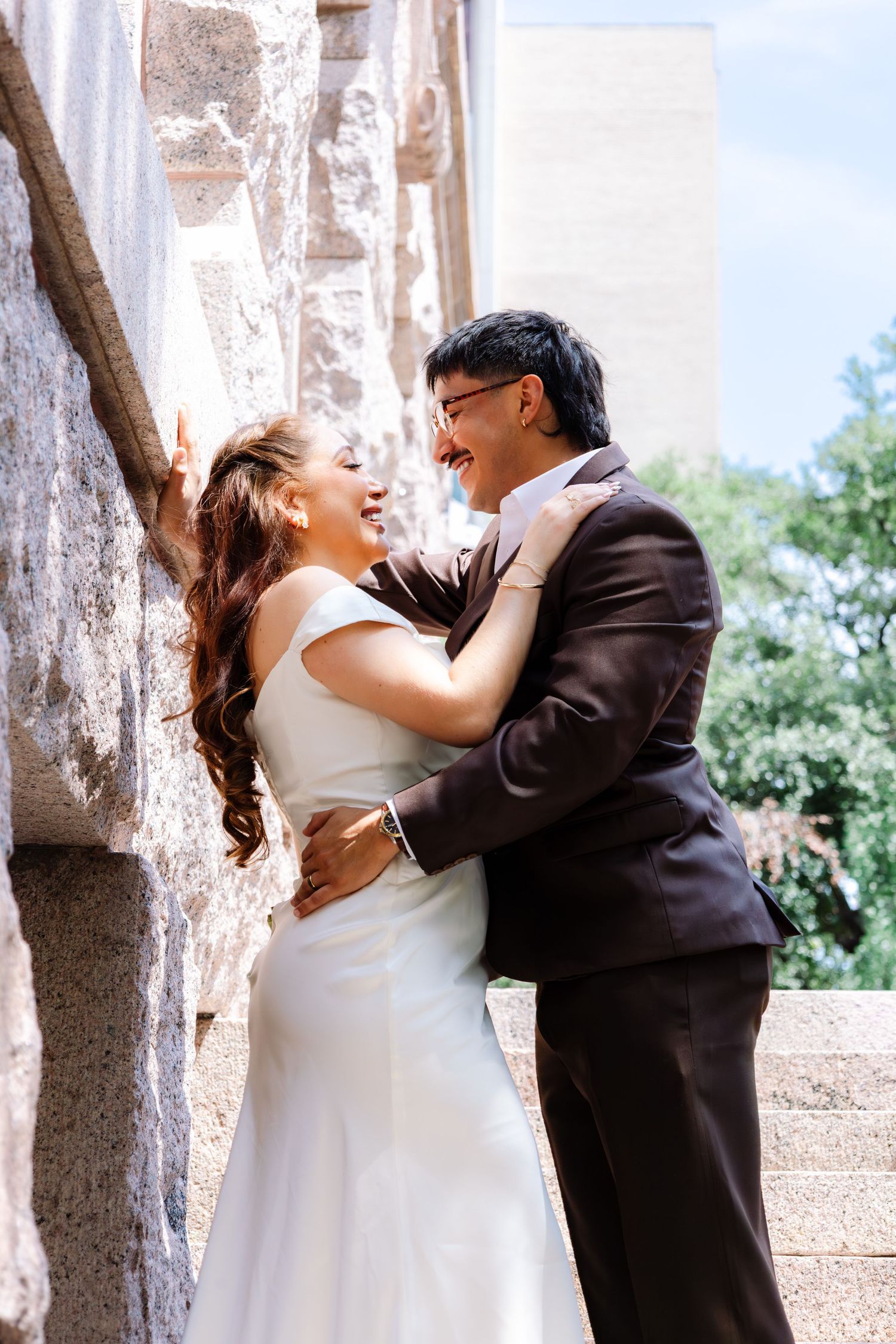 Smiling bride and groom embrace joyfully against a stone wall in outdoor wedding portraits.