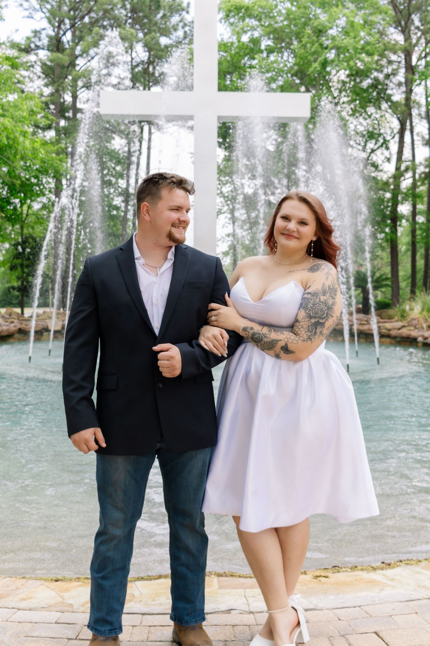 Couple posing before a white cross and fountain outdoors, woman wearing a white dress with tattoos.