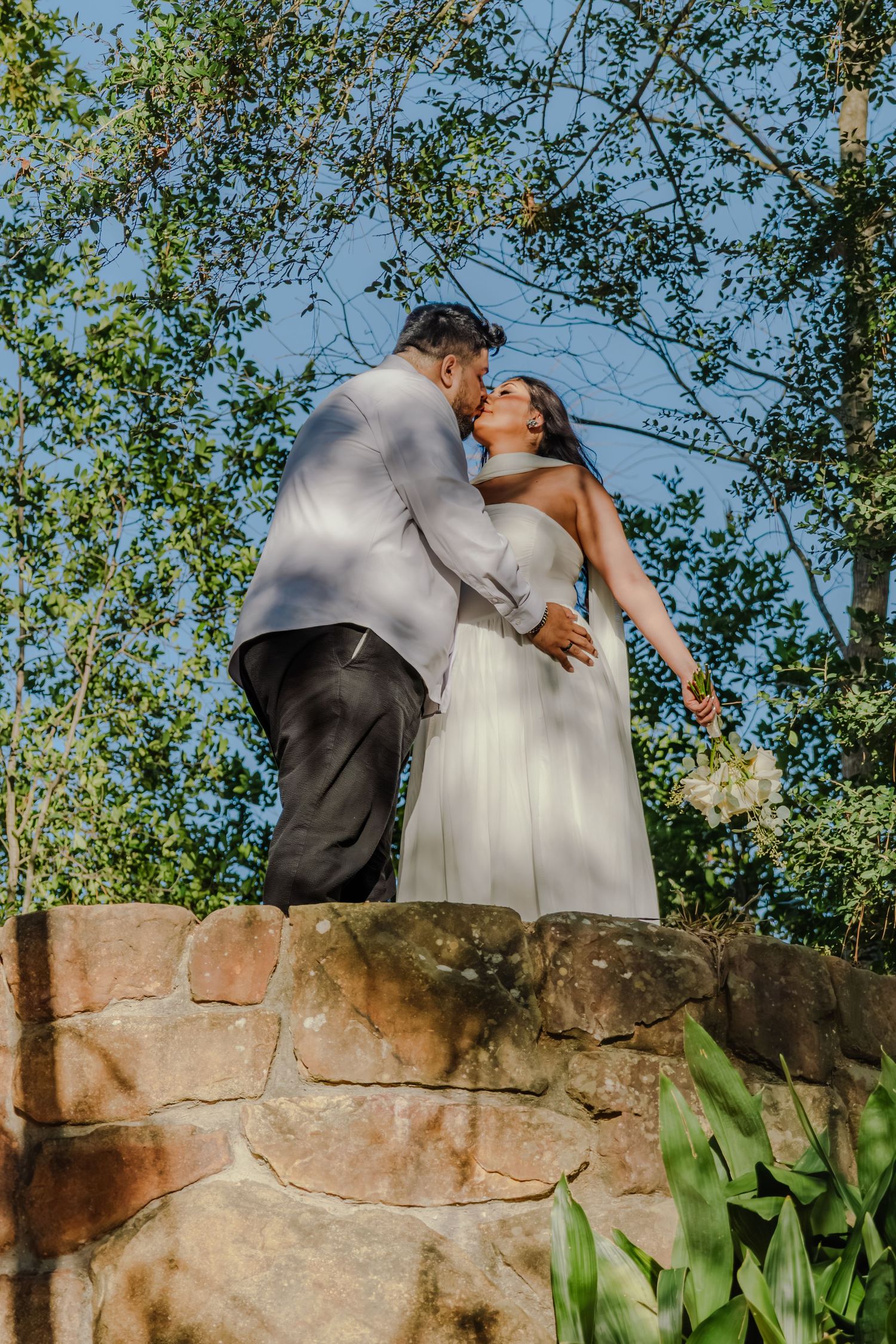 Bride and groom share a romantic kiss atop a stone wall surrounded by lush green trees and blue sky.