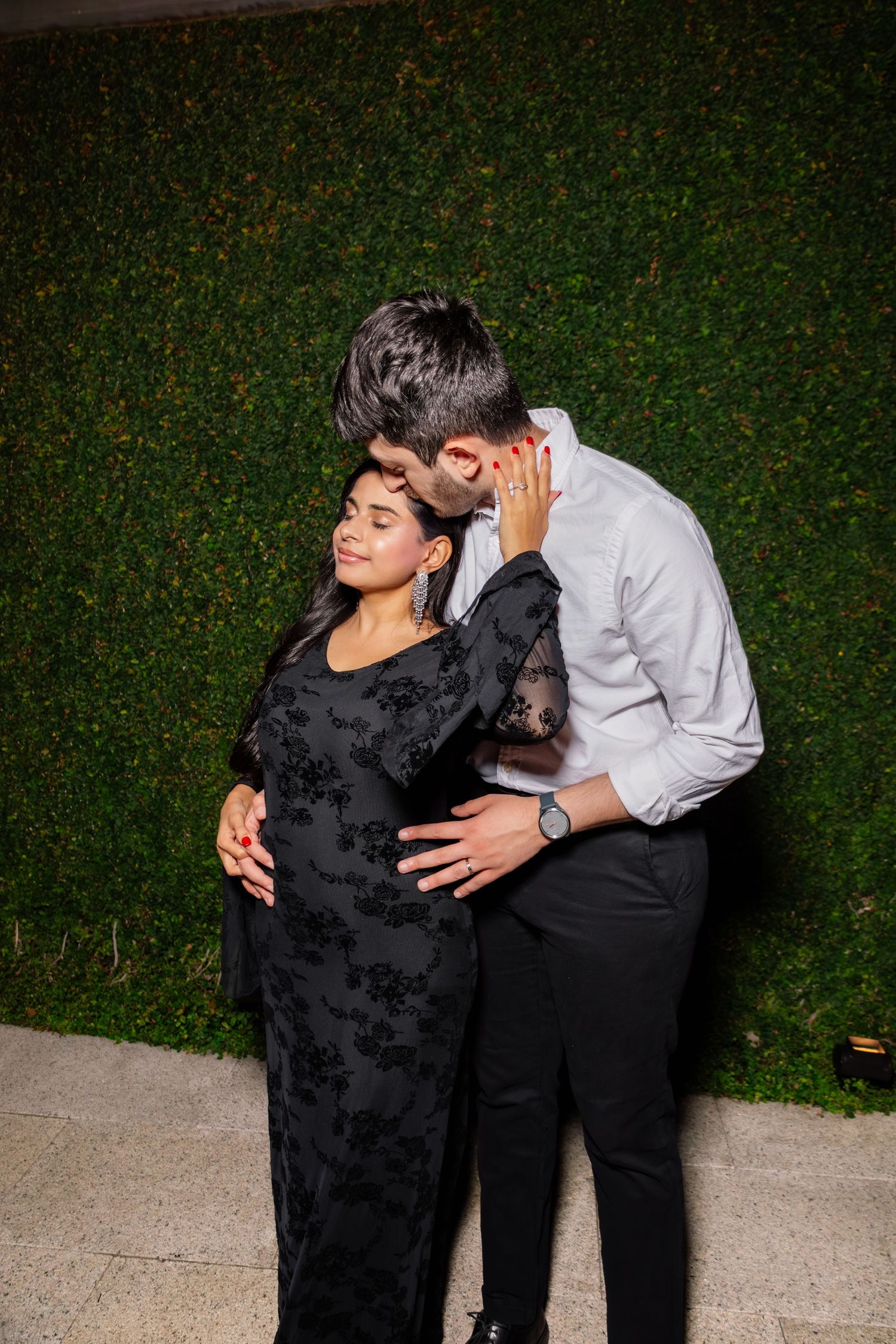 Couple embracing against green hedge wall at night, woman in black floral dress with man in white shirt kissing her head.