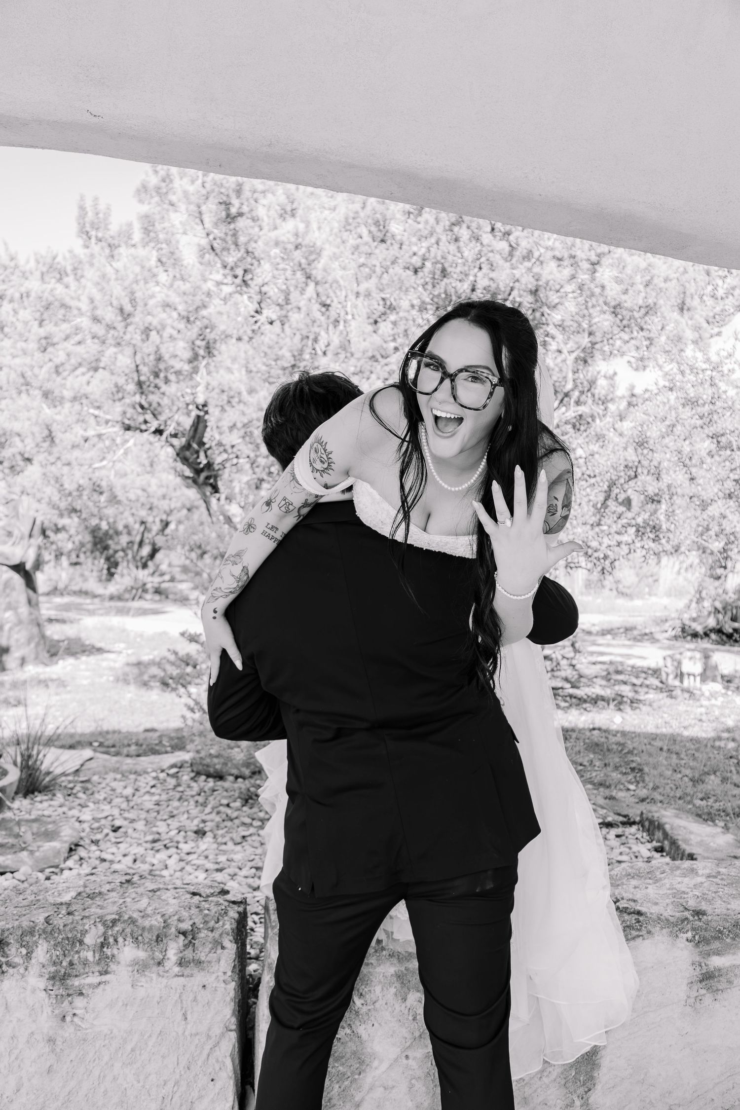 Black and white photo of a joyful bride in glasses showing her ring while groom embraces her outdoors.