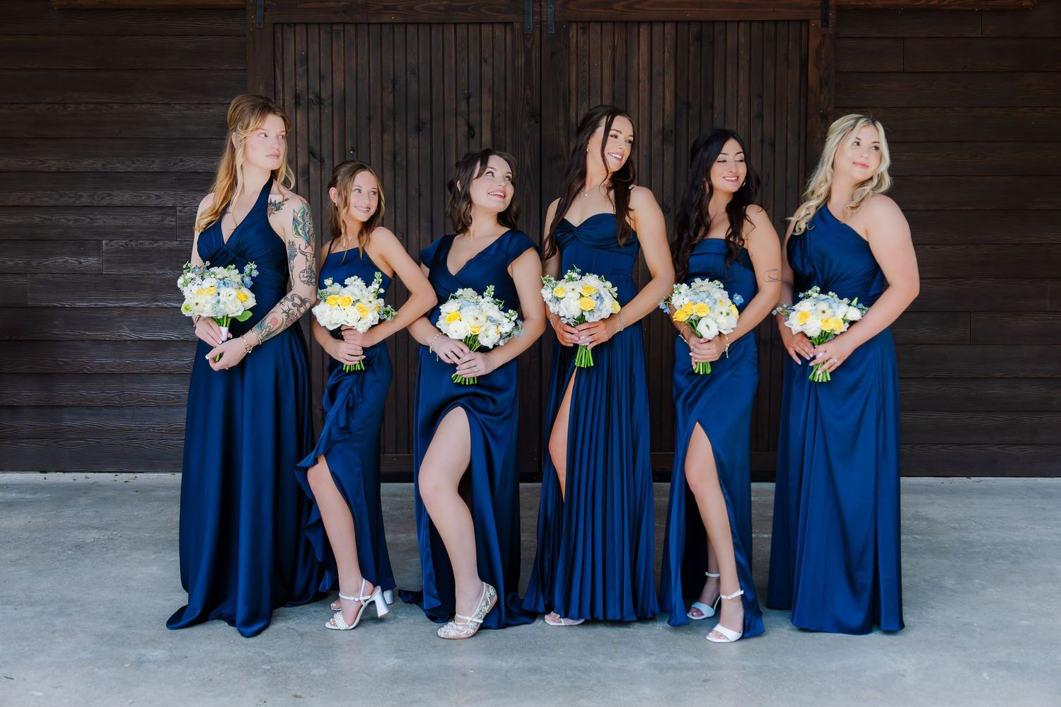 Six bridesmaids in navy blue gowns holding white bouquets pose elegantly in front of a rustic wooden barn wall.