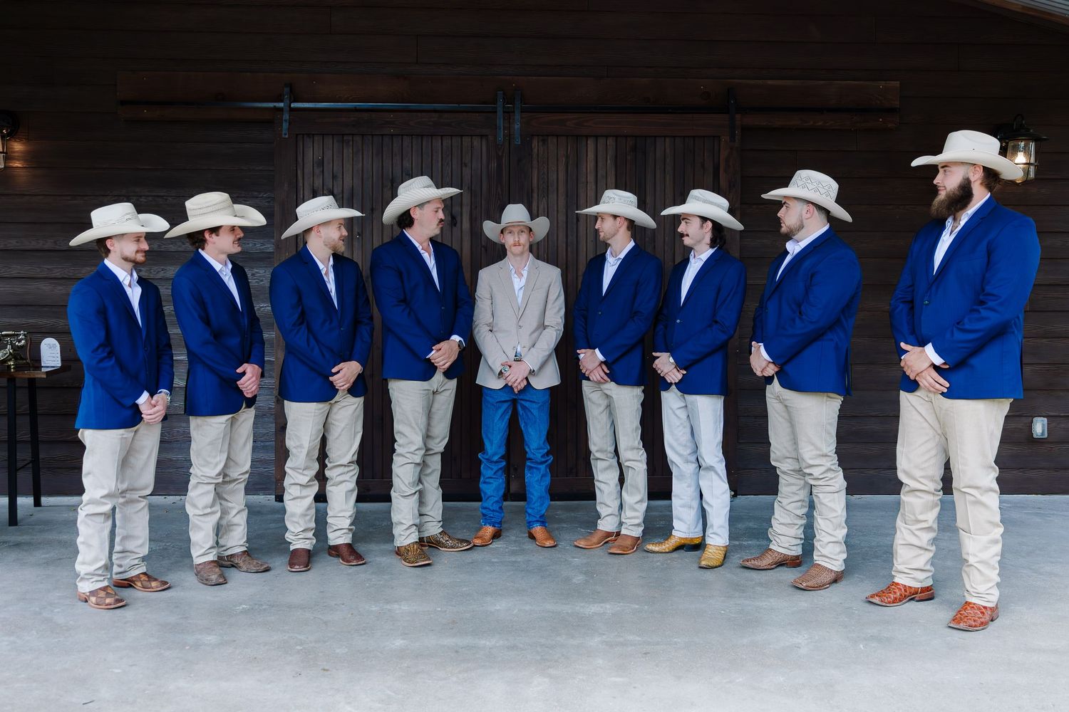 Groom in tan suit with groomsmen in blue blazers and cowboy hats posing outside a rustic barn wedding venue.