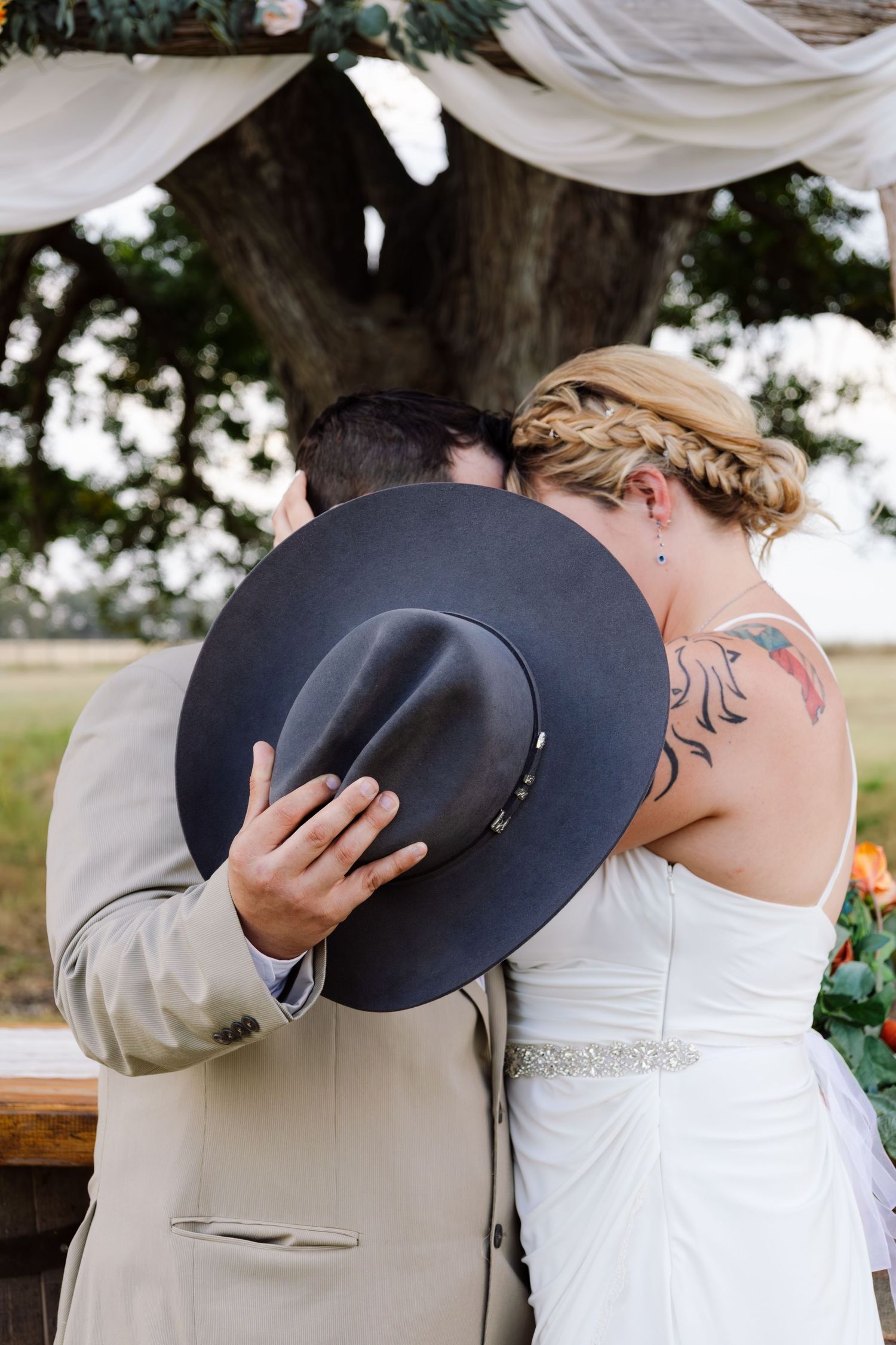 Bride and groom share a private kiss hidden behind a gray cowboy hat at their outdoor wedding ceremony.