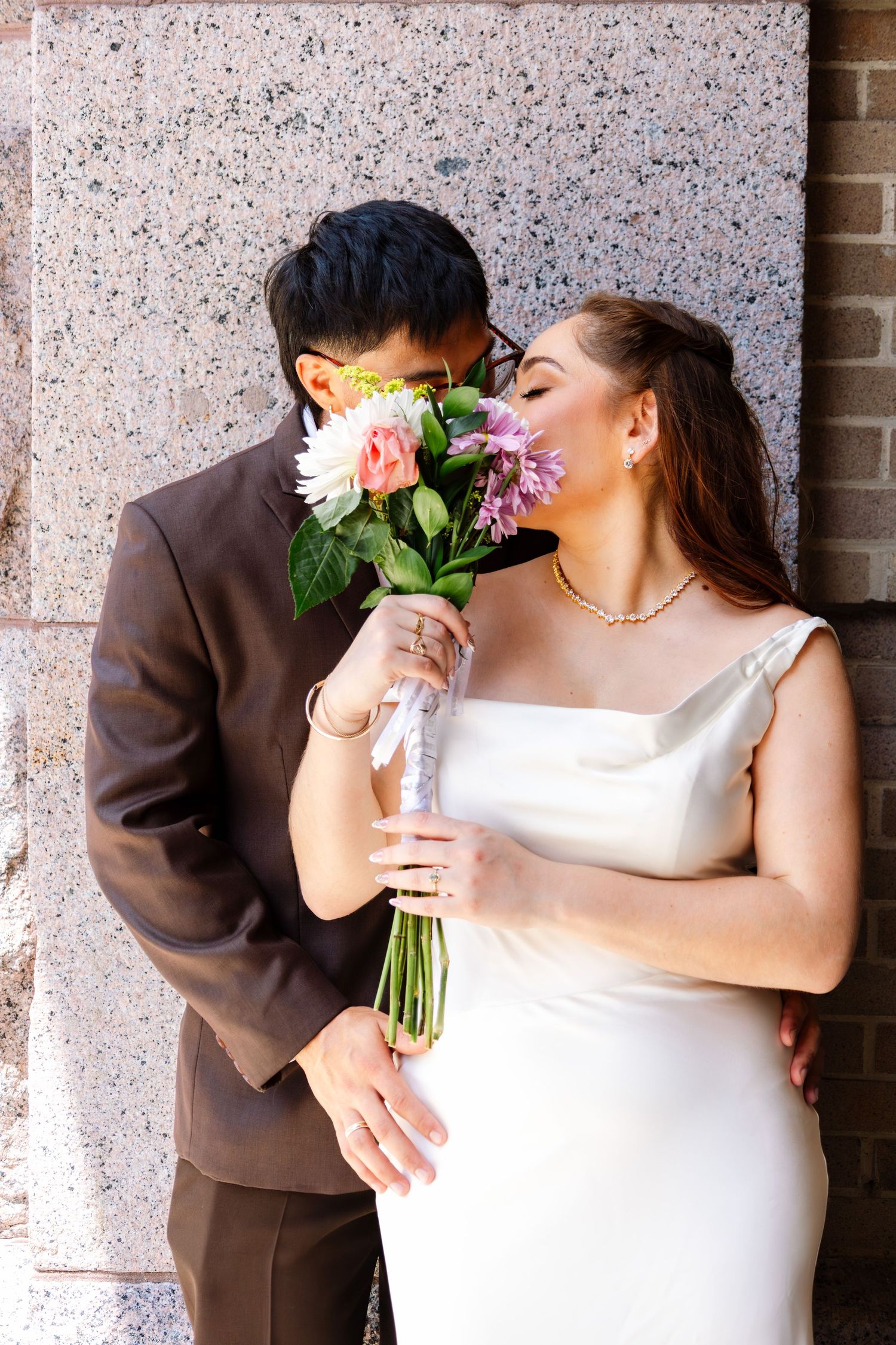 Bride and groom share a romantic moment hiding behind a colorful wedding bouquet outdoors.