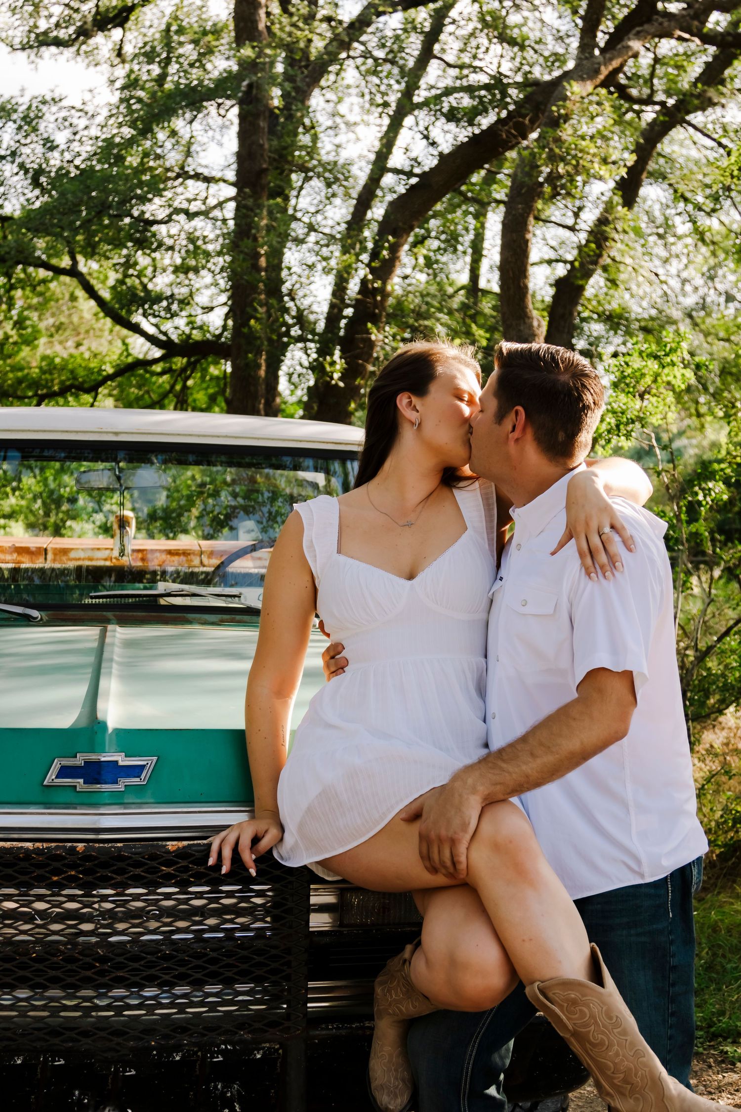 Couple sharing a romantic kiss leaning against a vintage teal Chevrolet truck under oak trees.