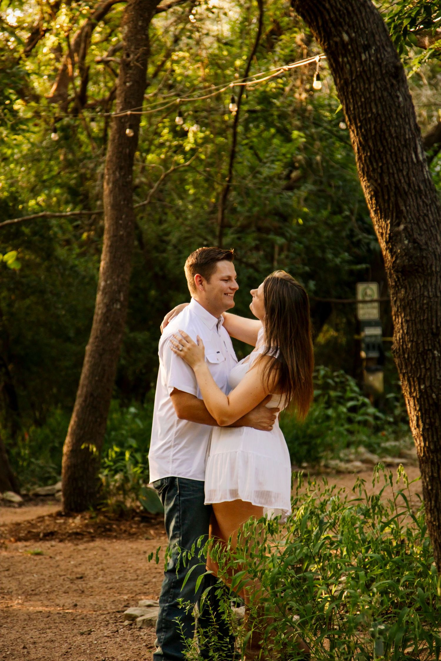 Smiling couple embracing on a sunlit forest path surrounded by lush green trees and foliage.