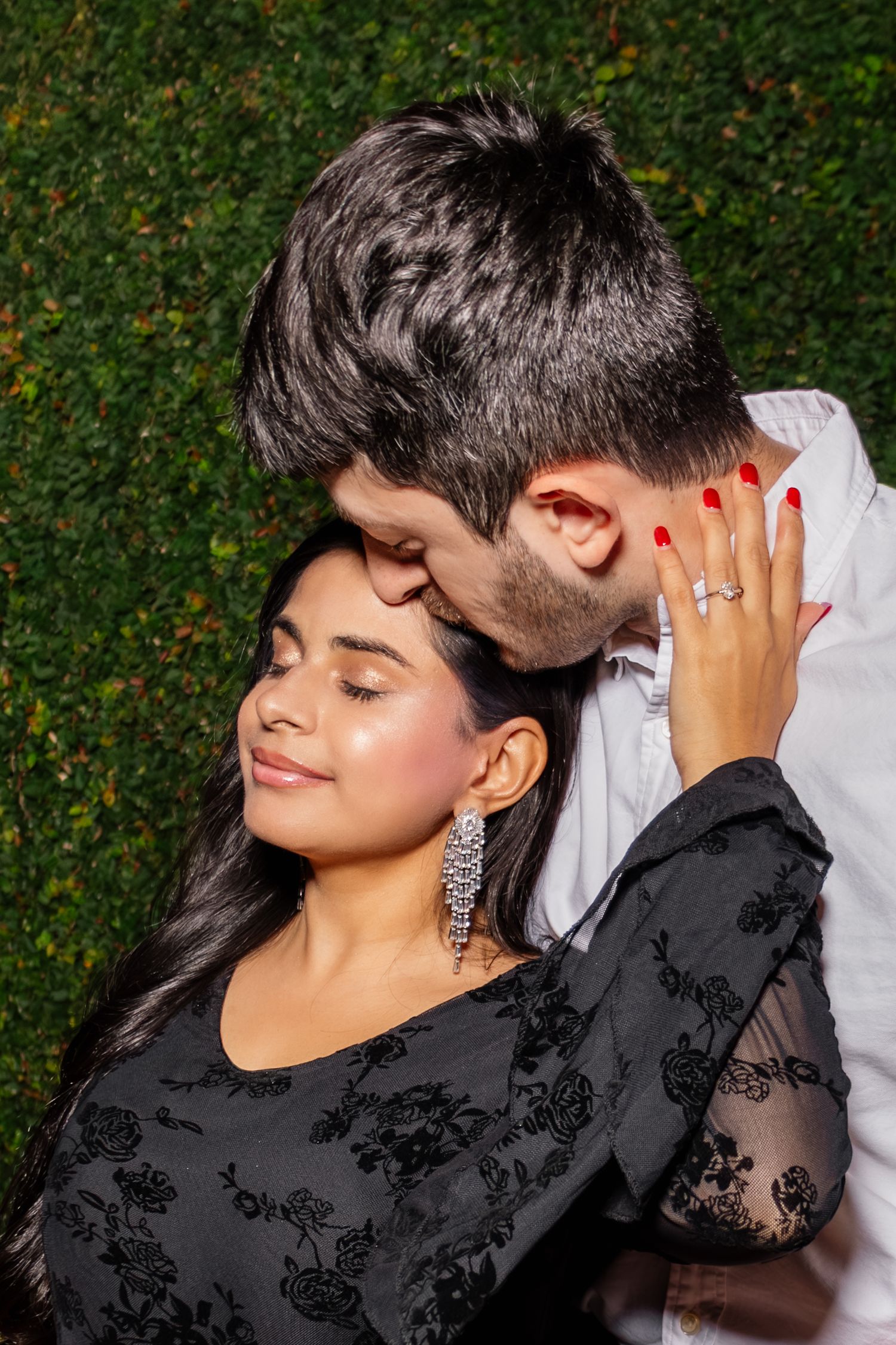 Man tenderly kisses woman's head as she displays her diamond engagement ring against a green hedge.