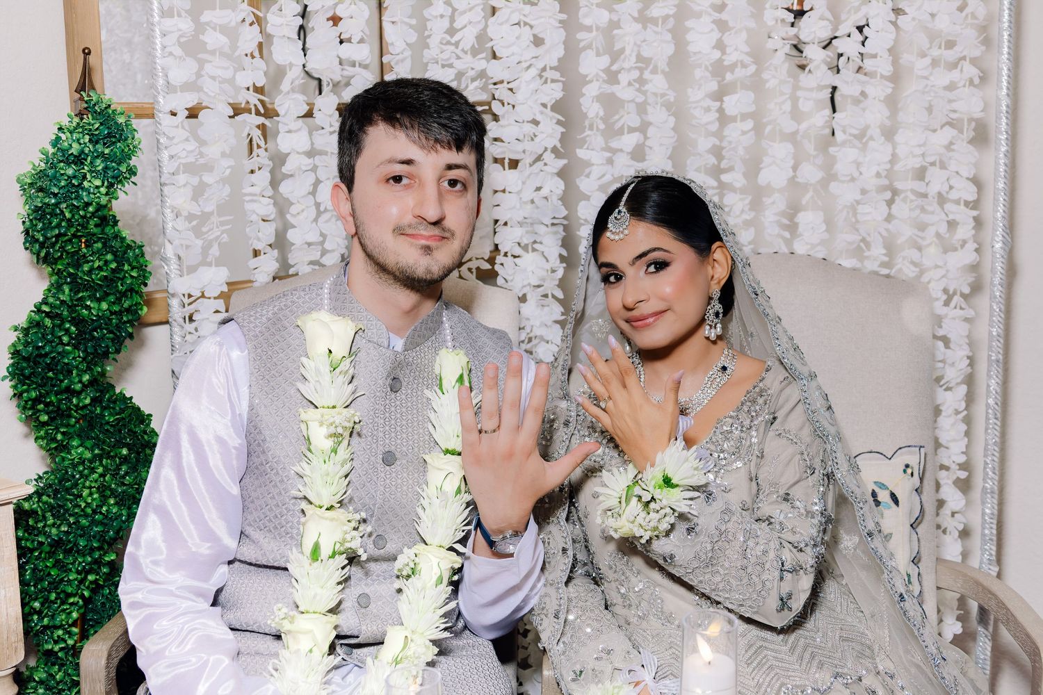 Newlywed couple in traditional South Asian wedding attire showing off their rings while seated on decorated bridal sofa.