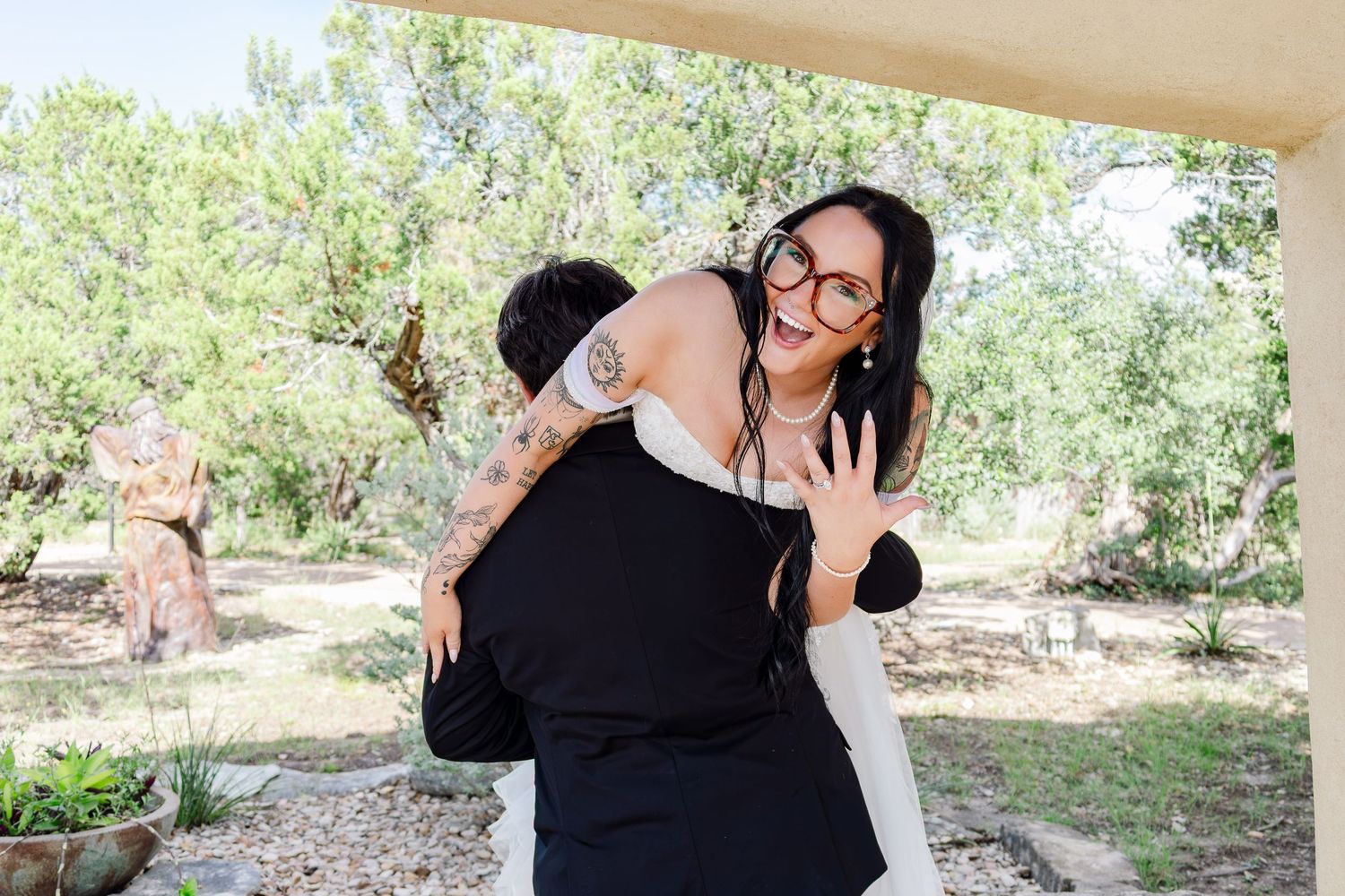 A joyful woman with glasses and tattoos laughs while being lifted in a hug outdoors near lush green trees.