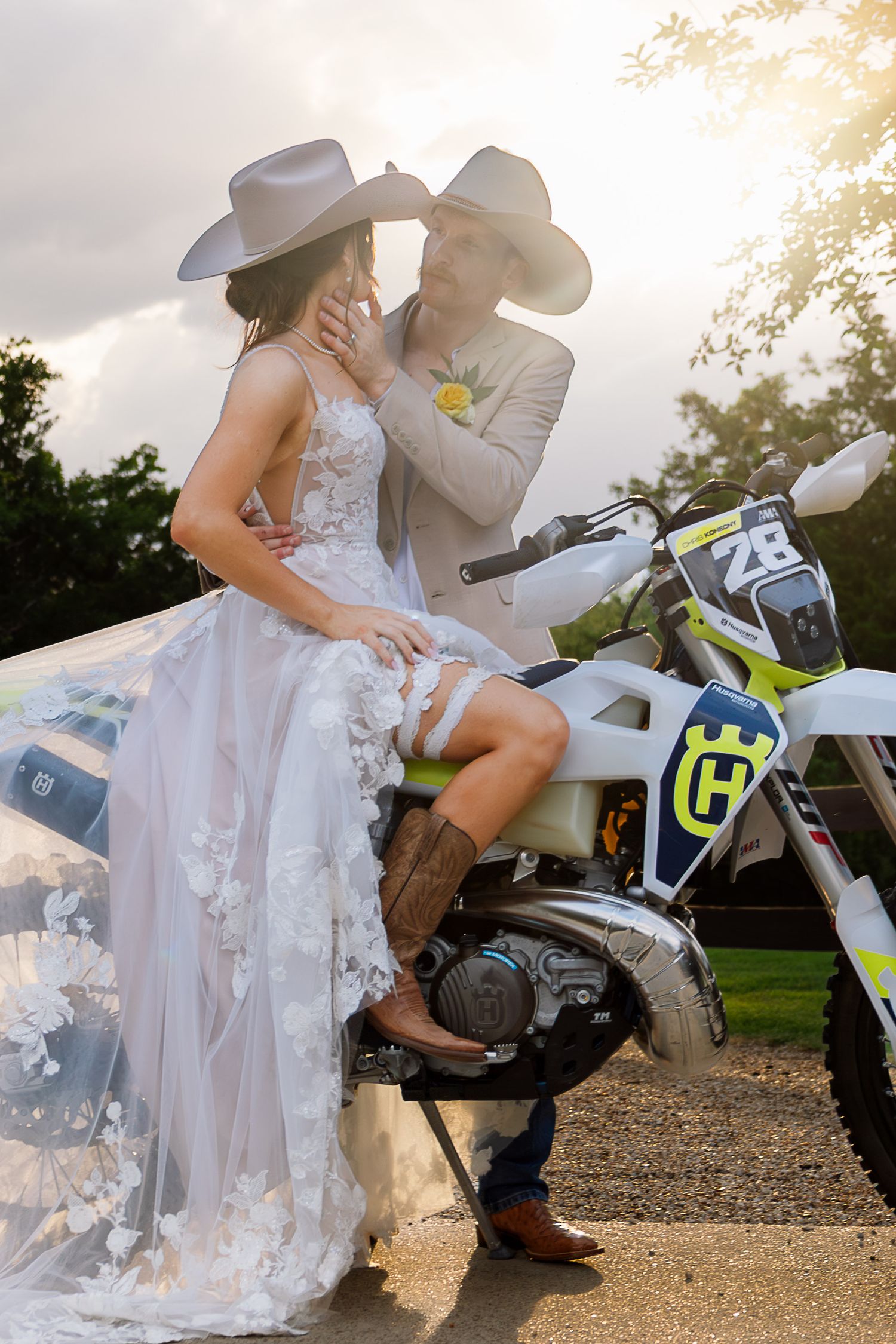 Bride in floral gown and cowboy hat sits on Husqvarna dirt bike with groom in tan suit at golden hour wedding.