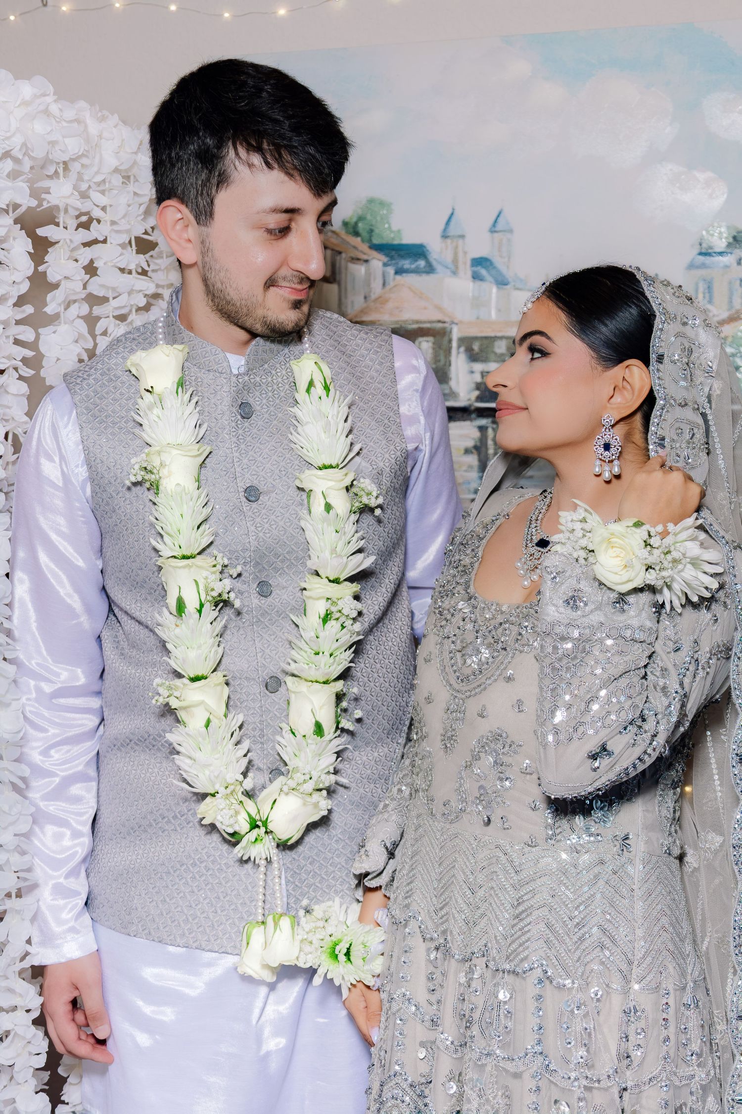 Couple in matching grey outfits with white floral garlands gazing at each other at a South Asian wedding ceremony.
