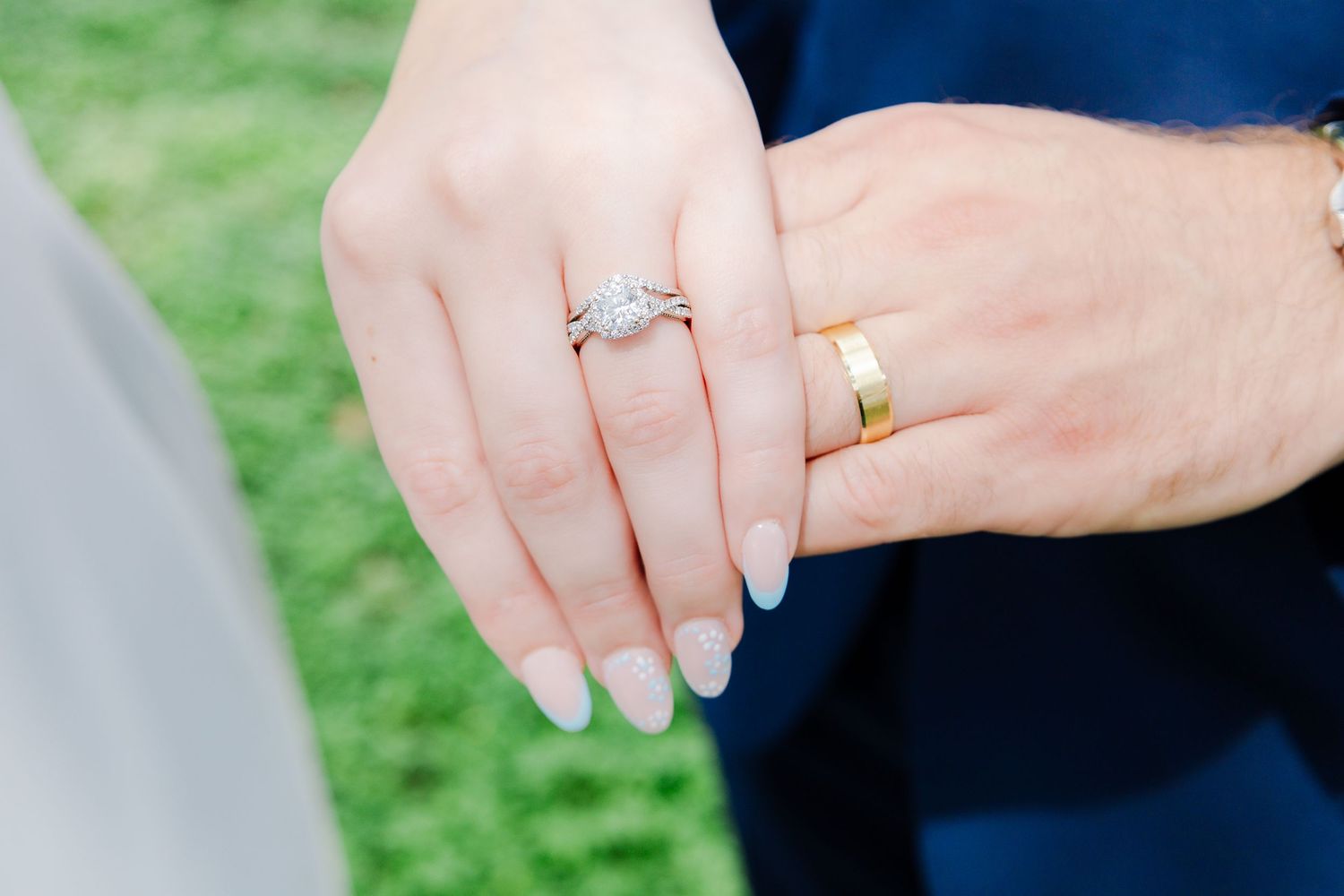 Close-up of bride and groom hands showing diamond engagement ring, wedding band, and gold wedding ring on green grass.