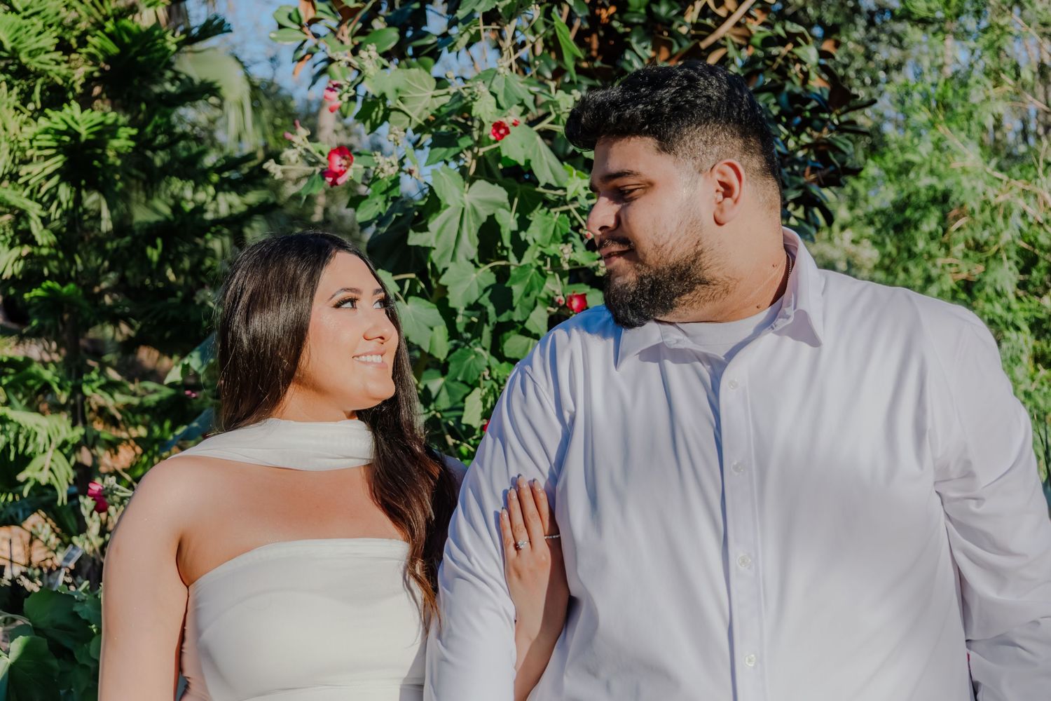 Couple smiling at each other outdoors with vibrant red flowers and green foliage in the background.